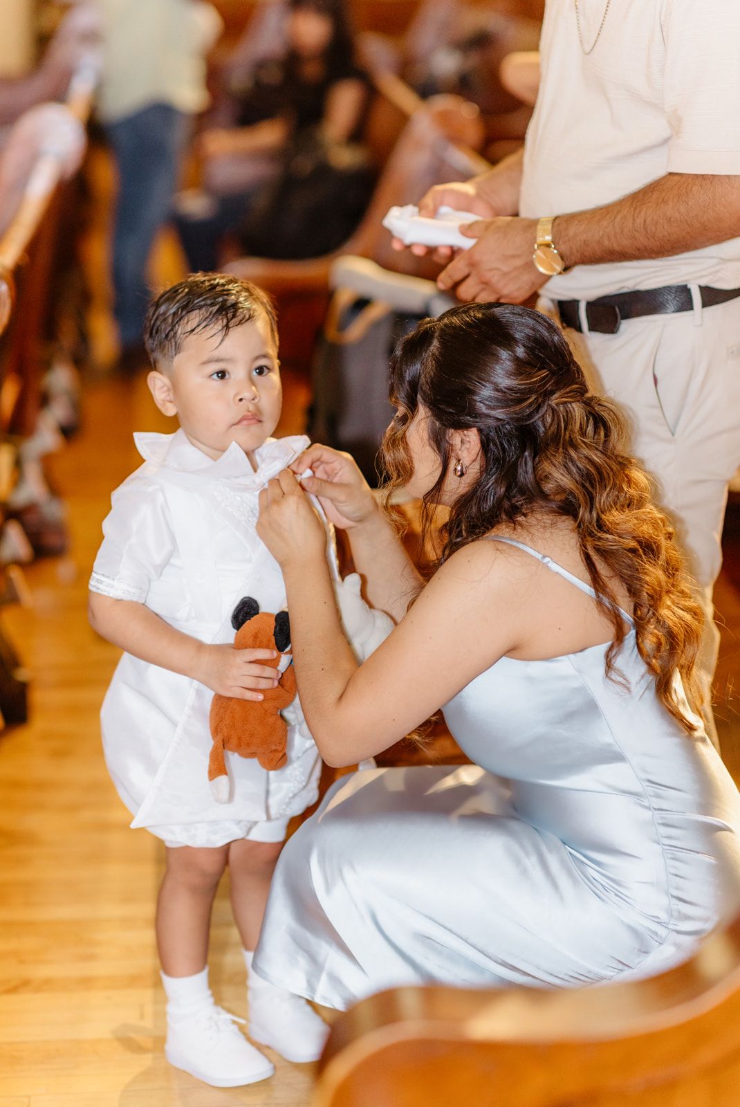 A woman in a blue dress adjusts a baby boys white baptism outfit in a church.