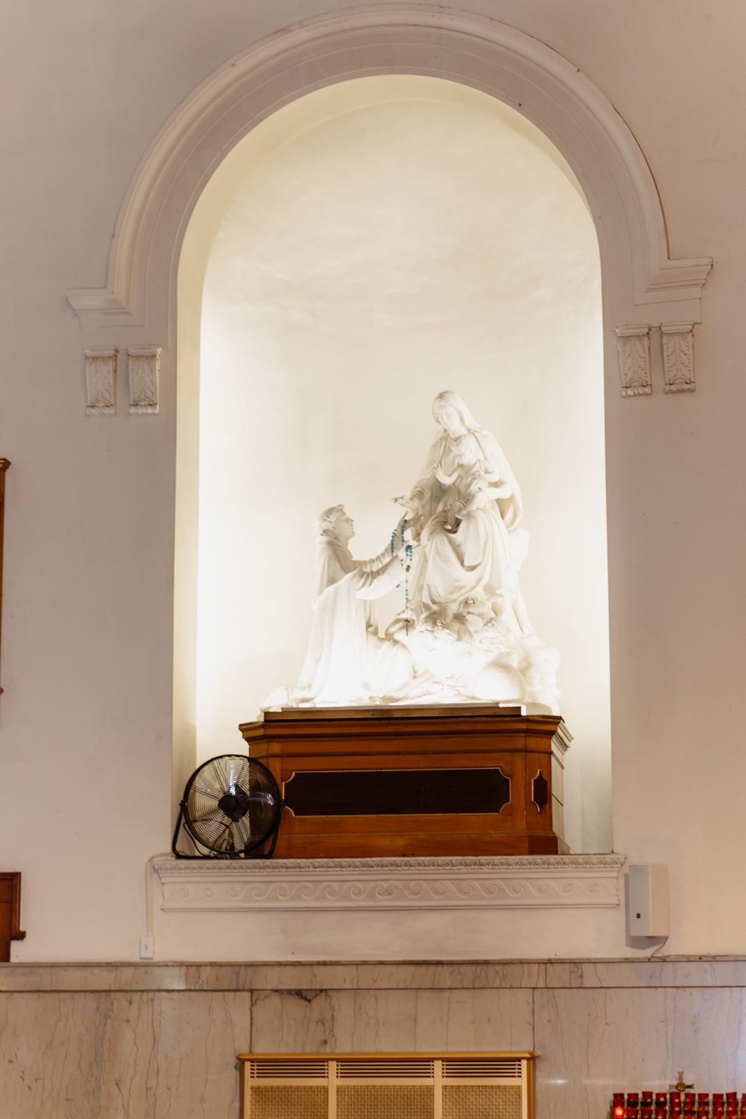 White marble religious statue displayed in an arched niche above a wooden pedestal inside a Catholic church.