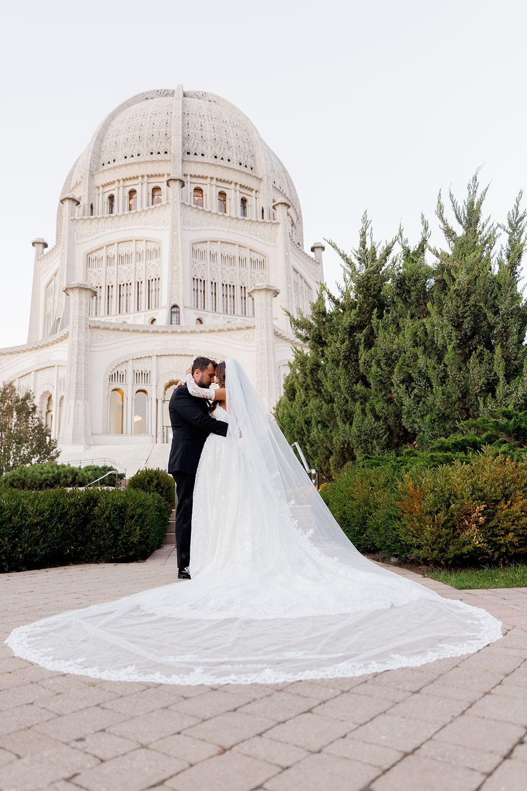 Bride and groom embrace in front of the Bahai Temple in Wilmette, Illinois, with a long cathedral veil trailing on the ground.