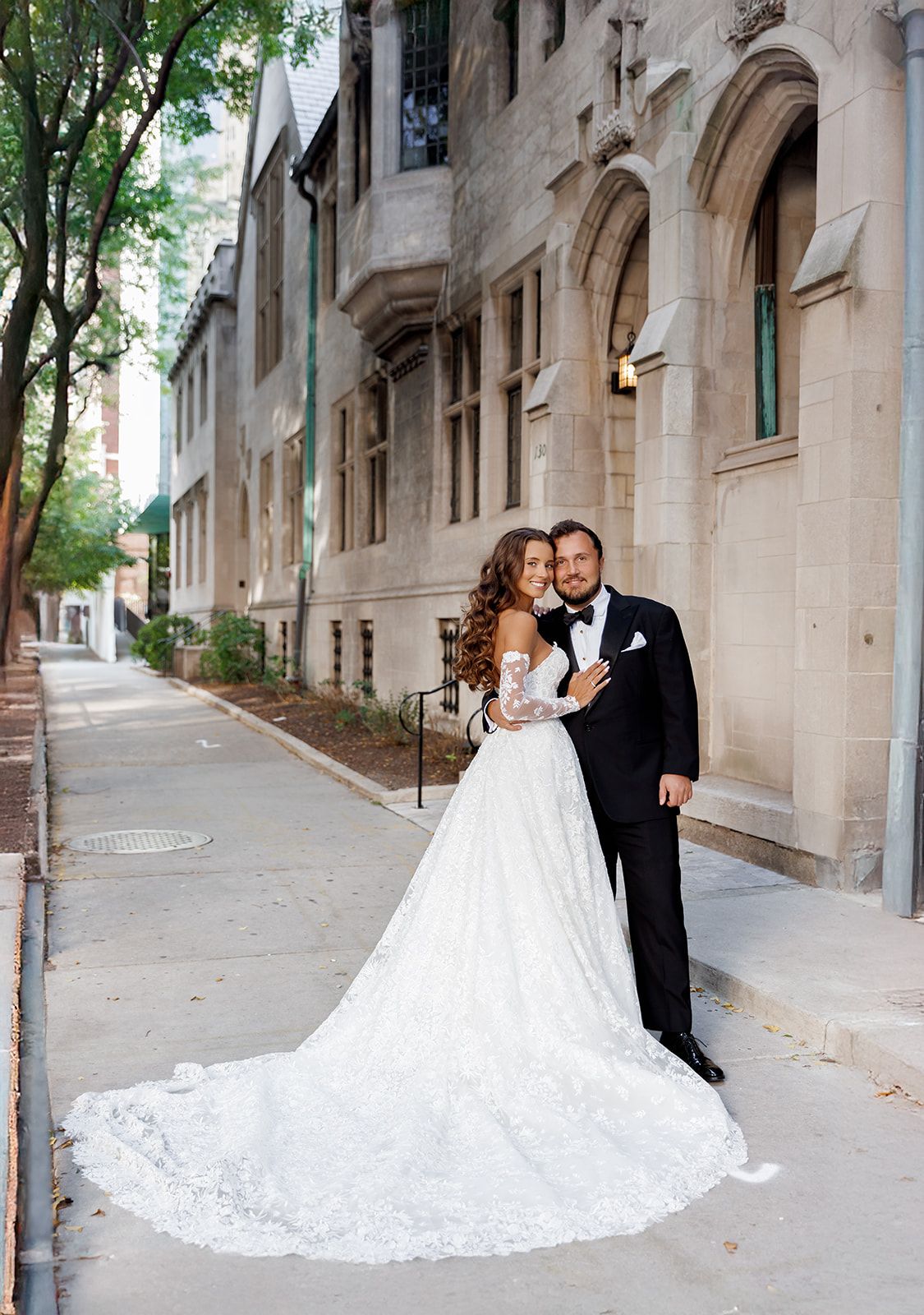 Bride in lace ball gown with cathedral train stands with tuxedo-clad groom on urban sidewalk.