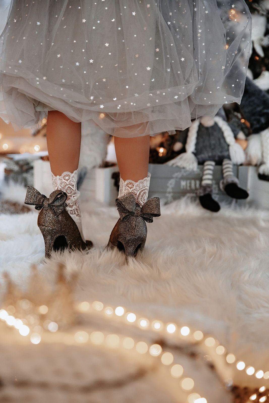 Child in silver tulle dress with lace socks and glittery bow shoes stands on white fur rug with Christmas lights.