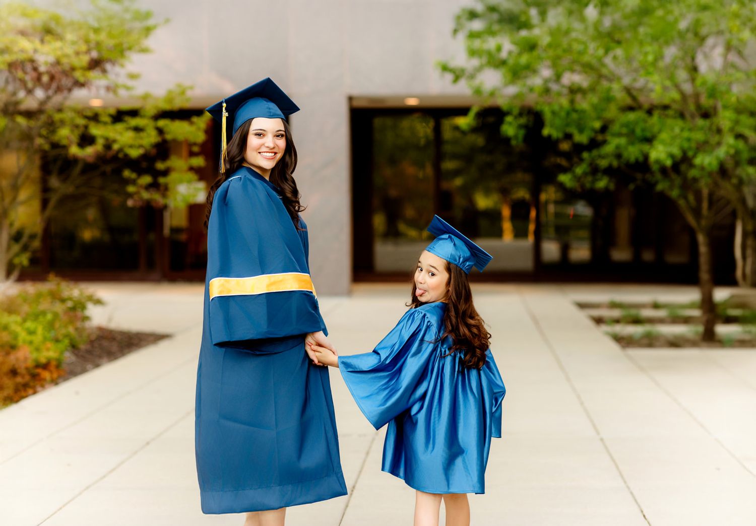 Graduate and young girl in matching blue caps and gowns holding hands on outdoor walkway.