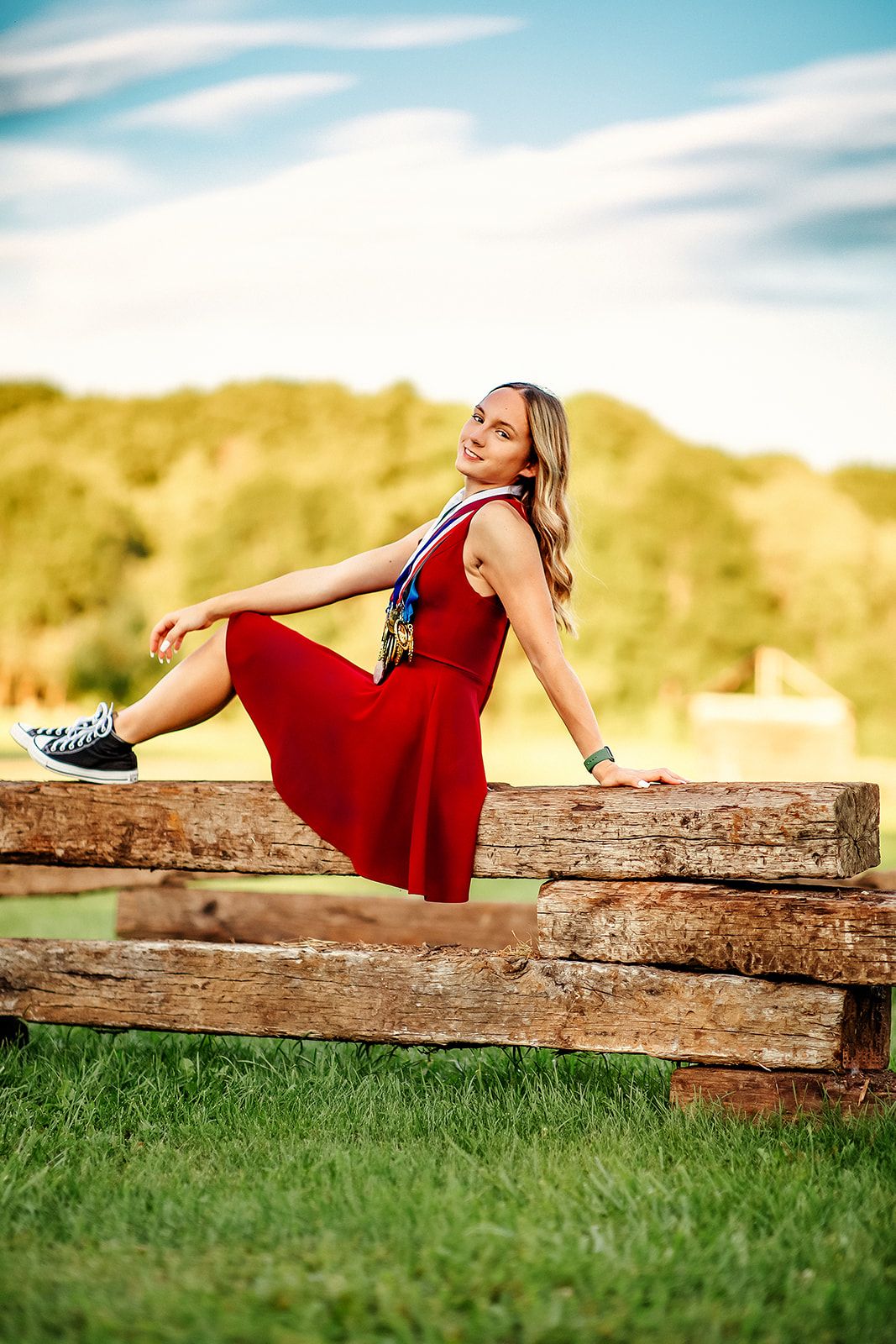 Smiling blonde teen in red dress sitting on wooden fence with medals outdoors.