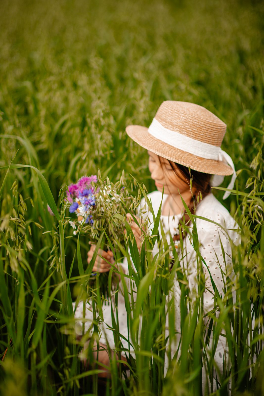 Woman in straw hat holding wildflowers while sitting in a lush green field.
