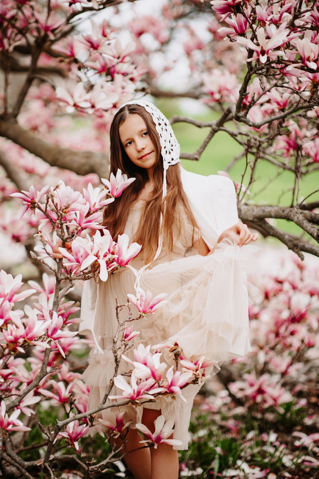 Young girl in white dress and bonnet posing among pink magnolia blossoms in spring garden.
