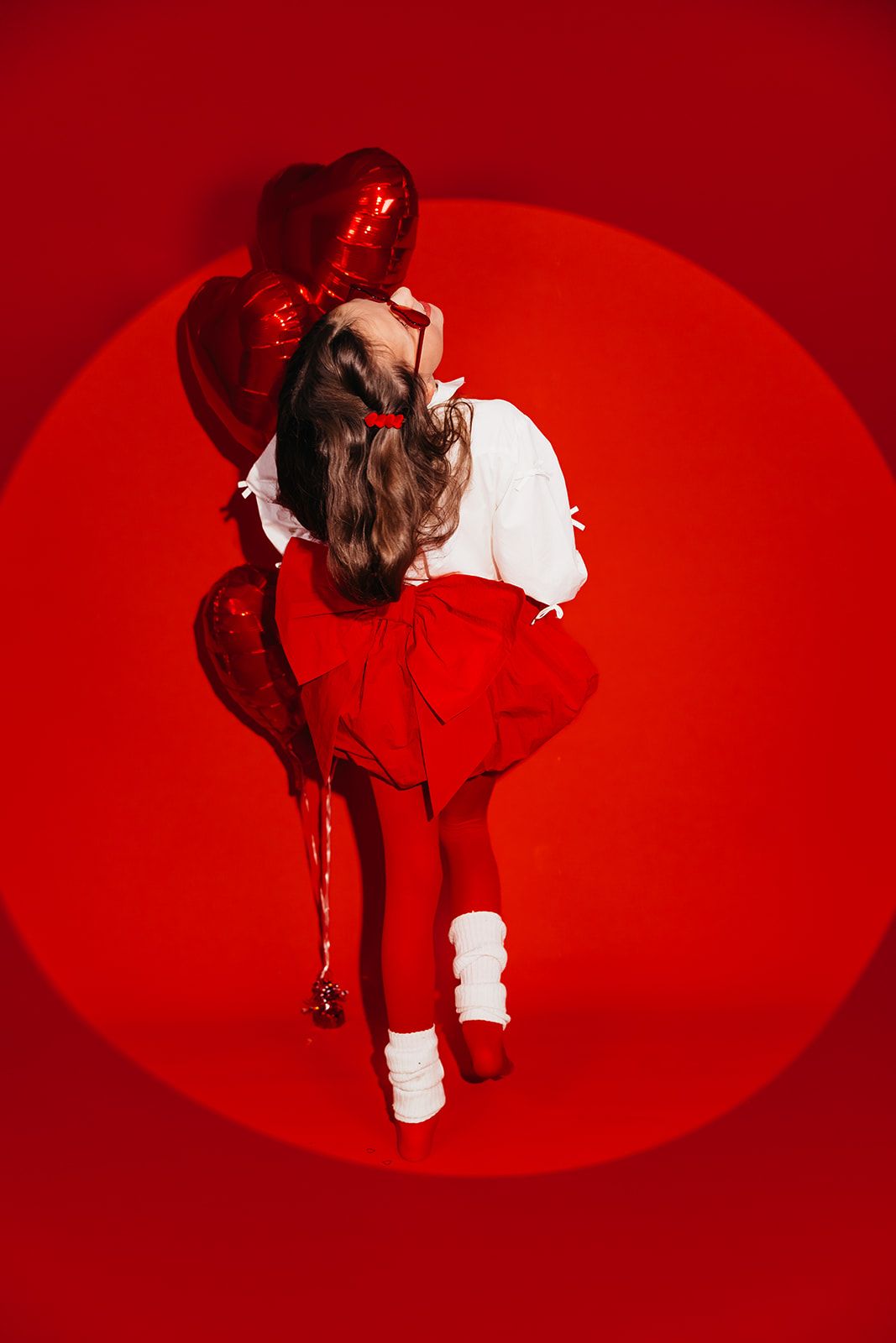 Young girl in red and white outfit holding red heart balloons on vibrant red background.