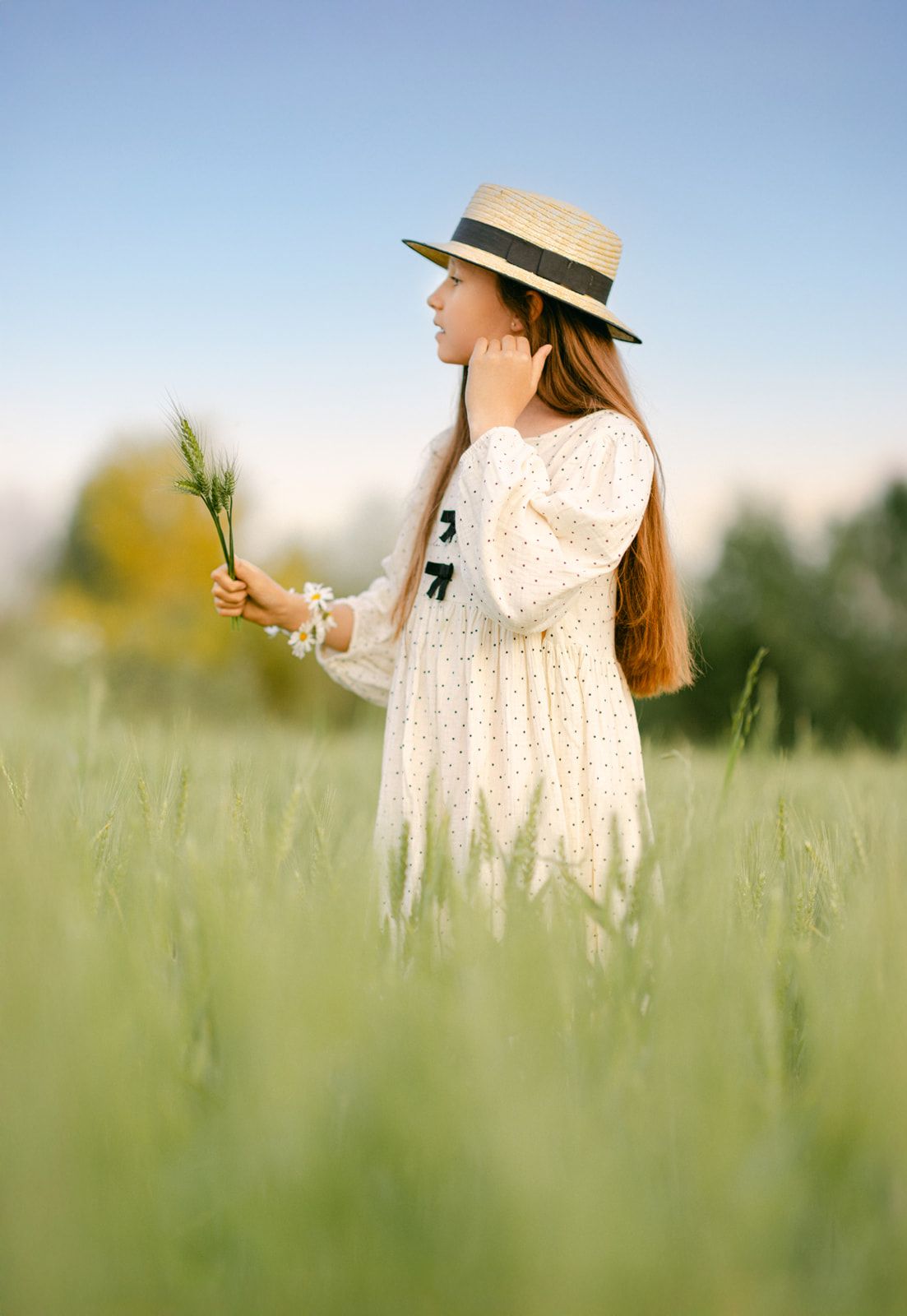 Young girl in straw hat and white floral dress holding wildflowers in a lush green field.