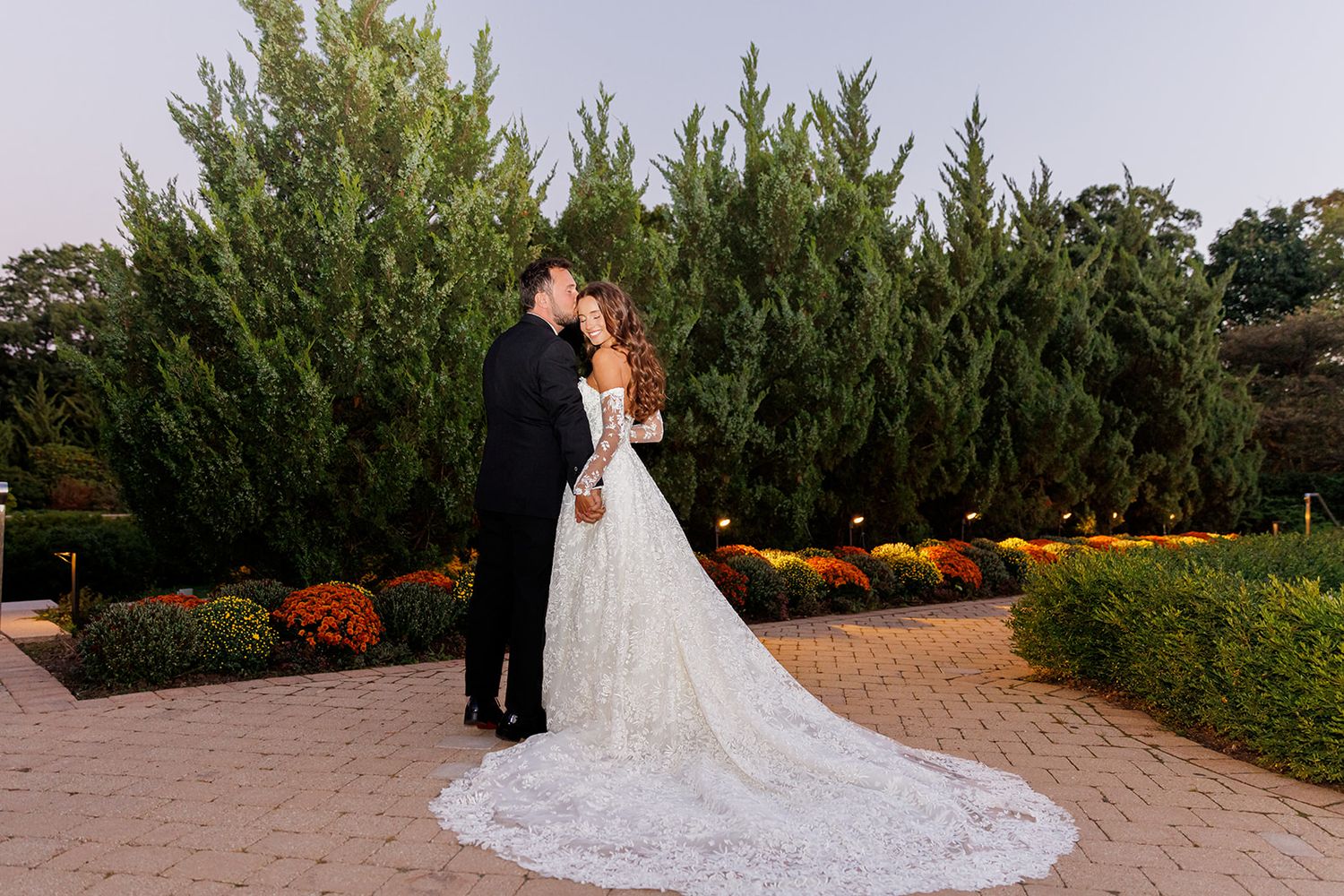 Bride and groom share a romantic kiss in a lush garden setting at dusk, bride wearing elegant lace gown with long train.