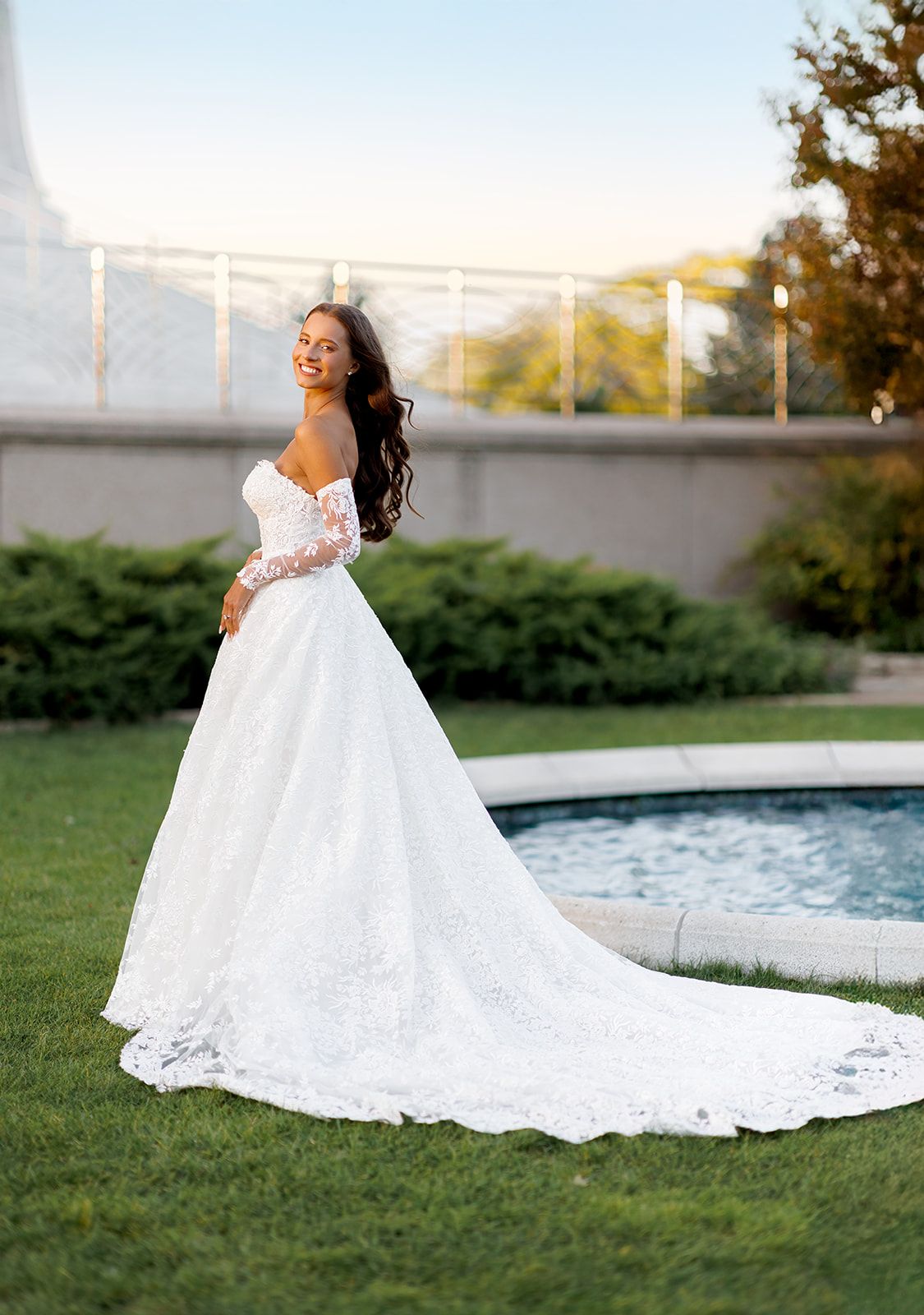 Bride in off-shoulder lace ball gown with long sleeves and cathedral train, posing outdoors near a fountain.