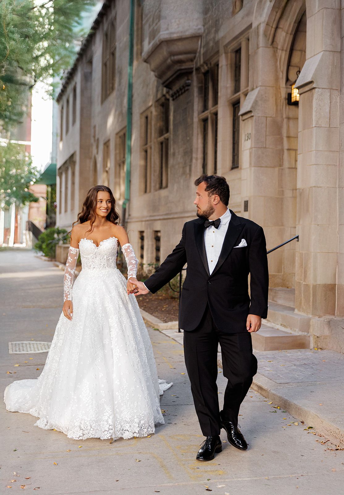 Bride in lace off-shoulder gown holds groom's hand in black tuxedo walking along historic stone building.