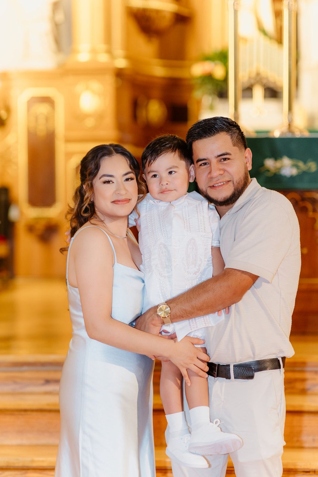 A young Hispanic family smiles together in a golden-lit church, cradling their child in a white baptism gown.