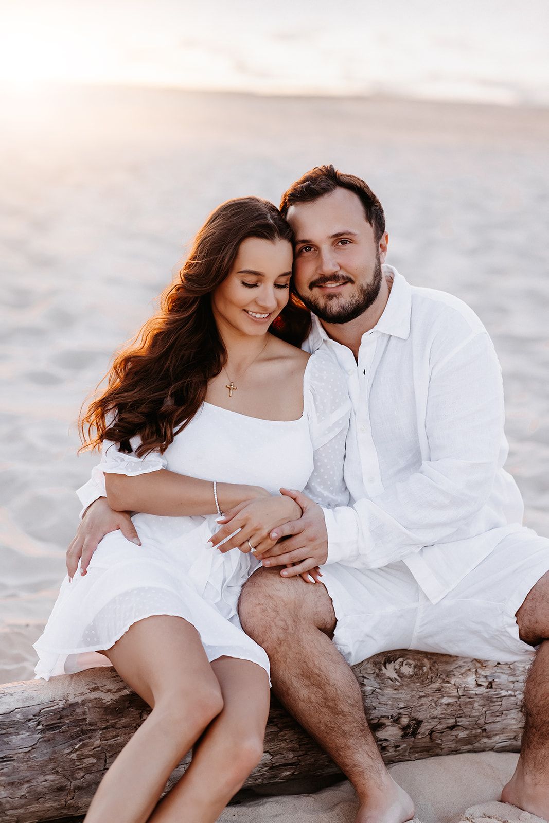 Romantic couple in white outfits sitting on driftwood at sunset beach, smiling and embracing.
