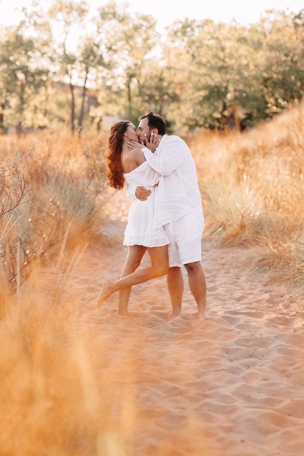 Couple kissing passionately on sandy path surrounded by golden grass and trees at sunset.