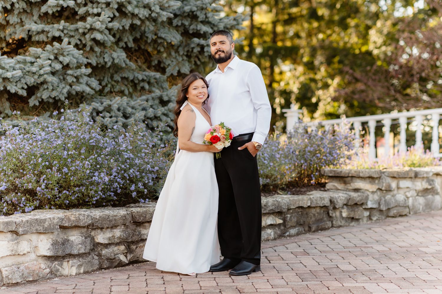 Bride and groom posing outdoors on brick path with lush greenery and white railing backdrop on their wedding day.