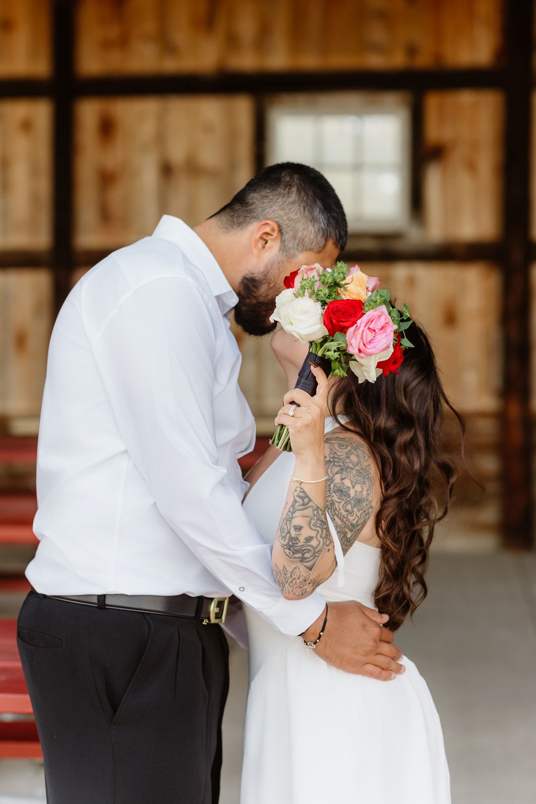 Bride and groom sharing kiss hidden behind colorful rose bouquet inside wooden barn.