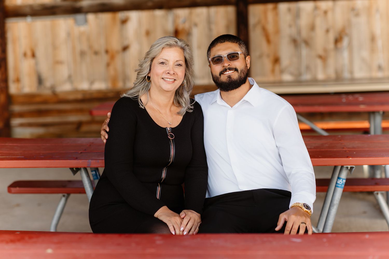 A smiling couple poses together at a rustic barn venue, sitting at a red picnic table.