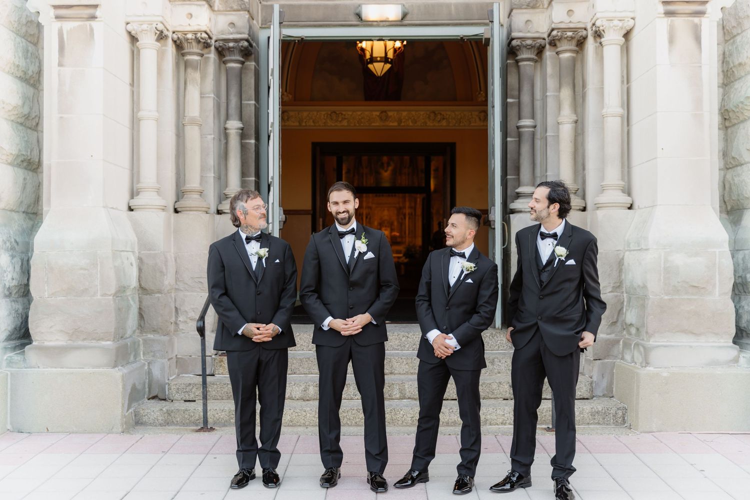 Four groomsmen in black tuxedos posing together outside elegant stone building before wedding.