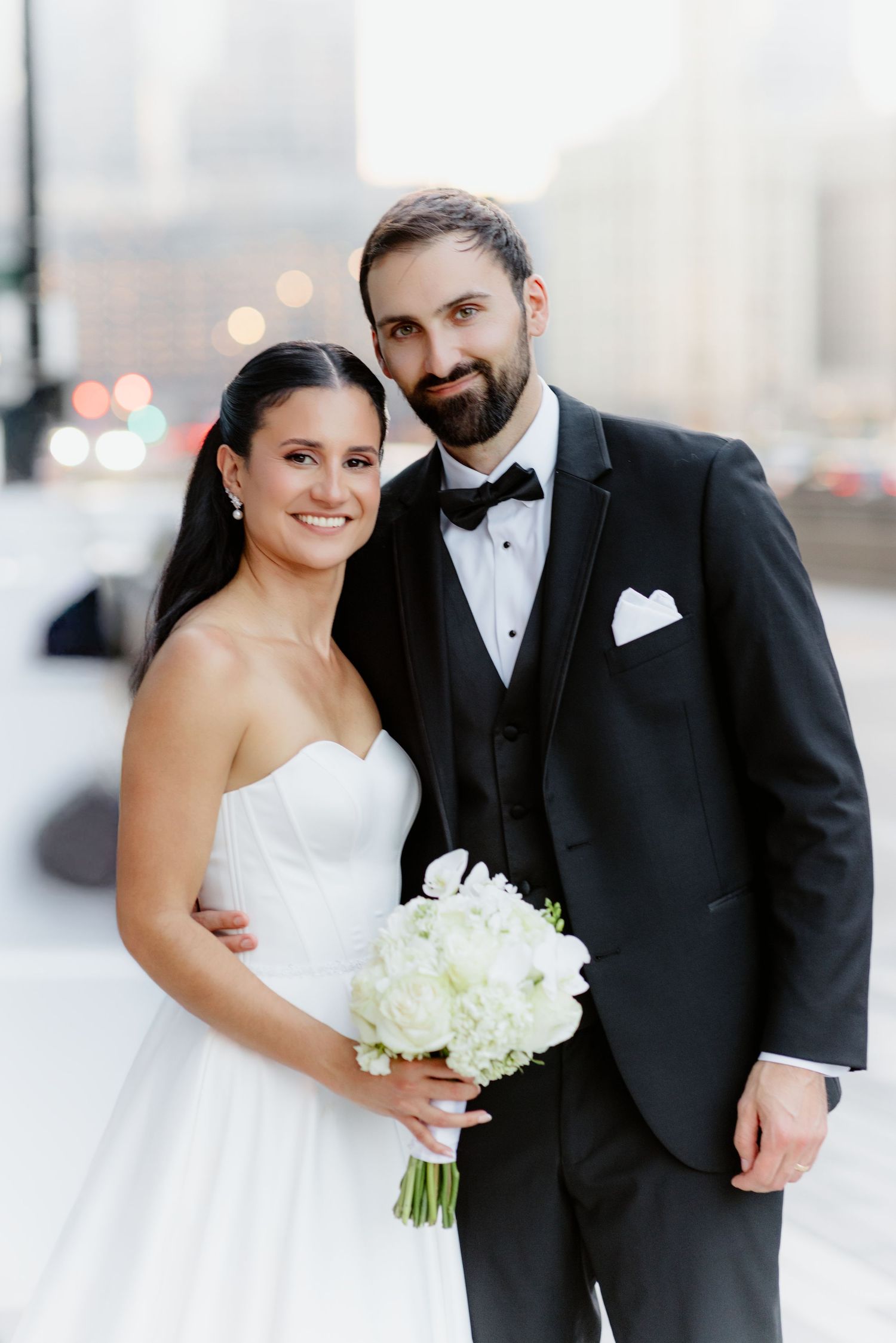 Smiling bride and groom in black tuxedo pose together with white bouquet against a blurred city backdrop.