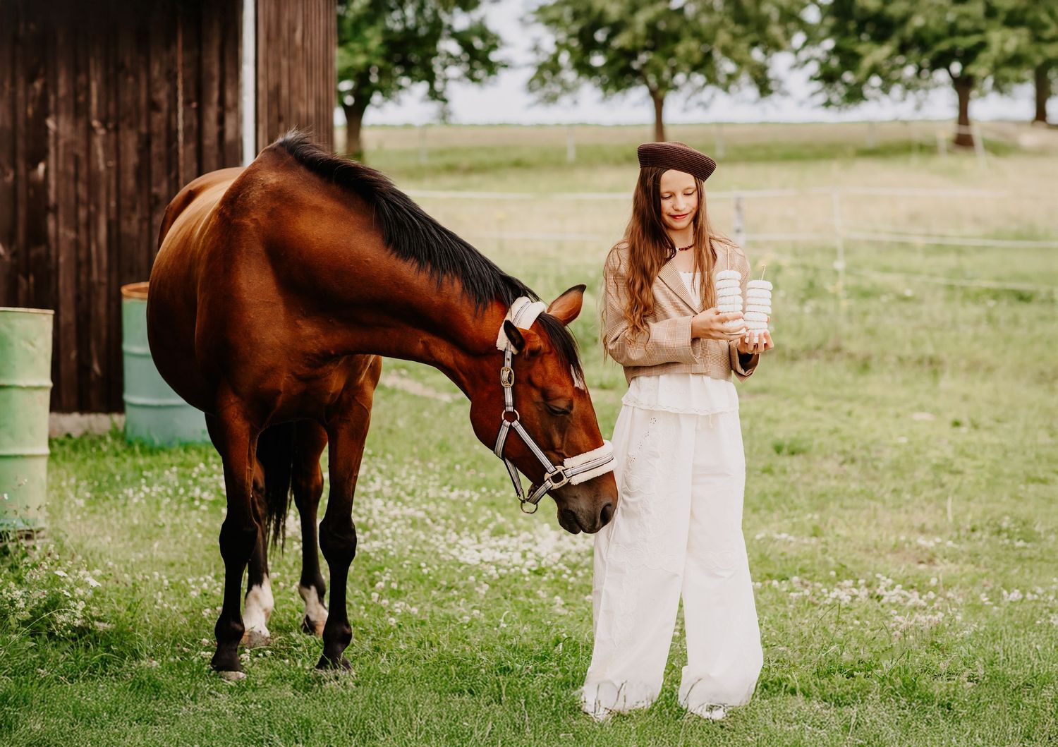 Woman in white outfit offers a treat to a chestnut horse near a rustic barn on a farm.