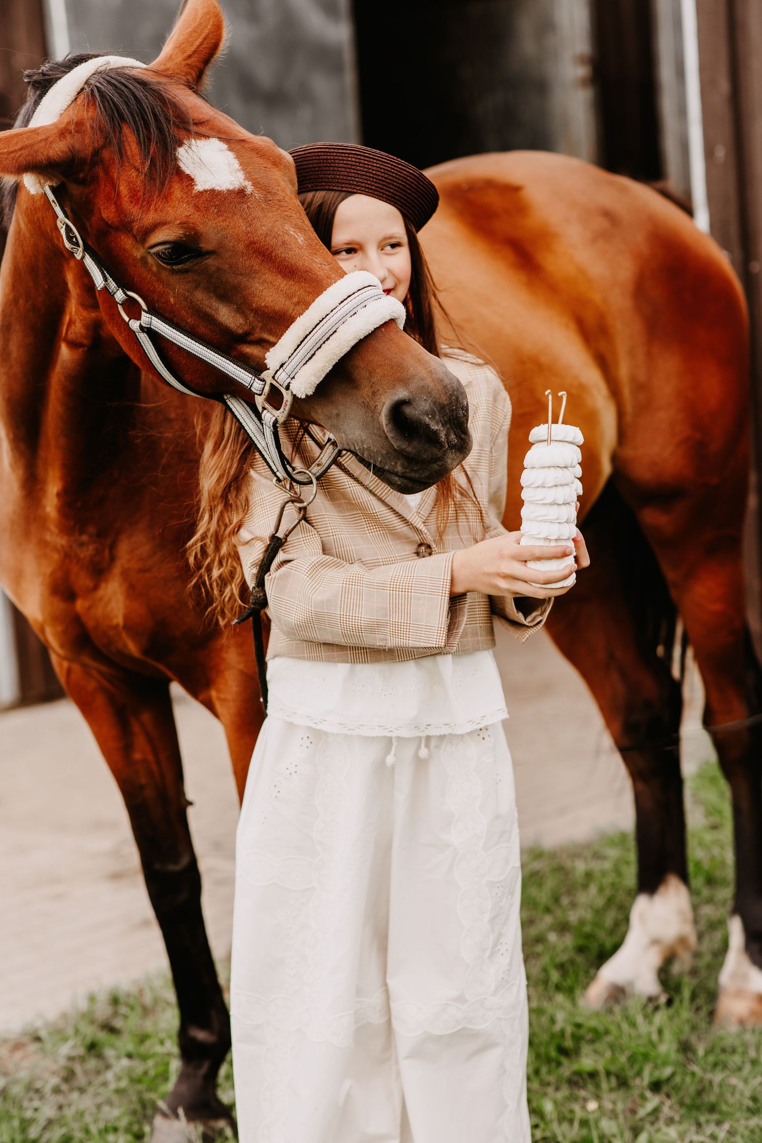 Young girl in brown hat and beige outfit standing close to a chestnut horse while holding a white treat stack.