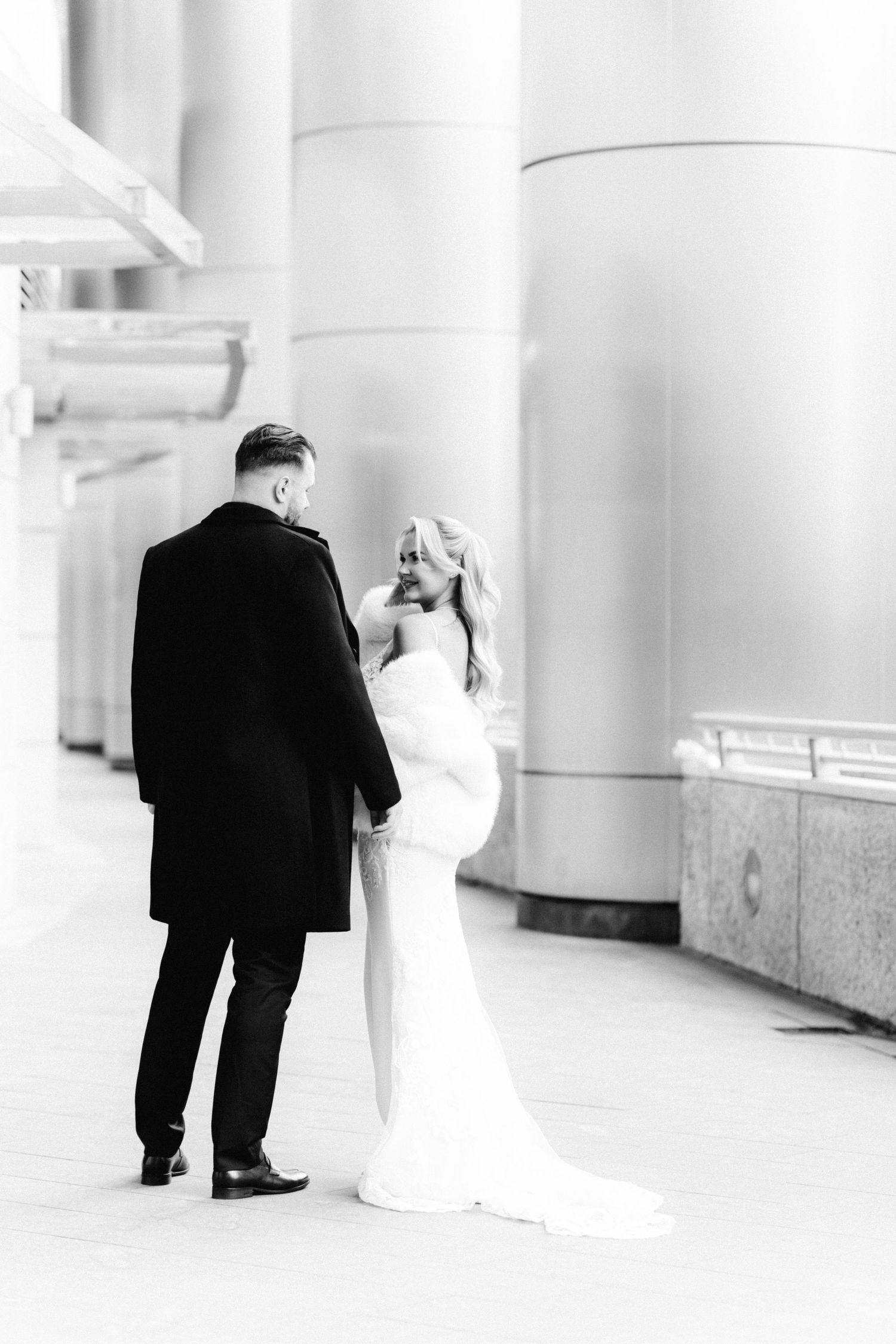 Black and white photo of bride and groom holding hands among large silver columns downtown.