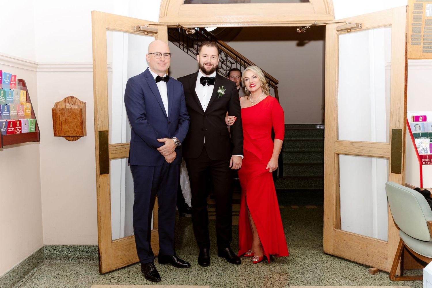 Groom in black tuxedo stands between parents in blue suit and red dress at wedding venue entrance.