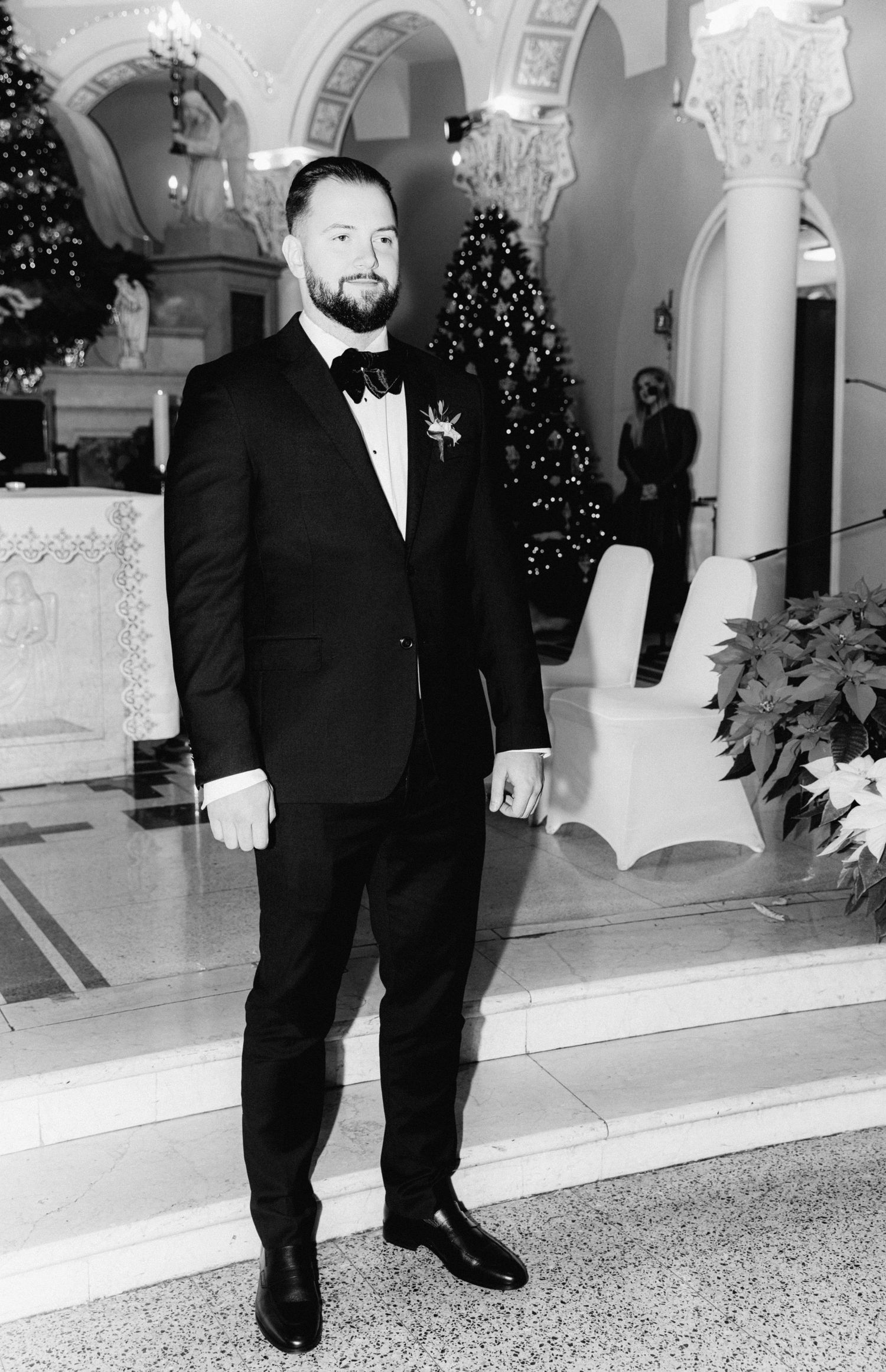 A groom in a black tuxedo poses in black and white before a Christmas tree at the church altar.