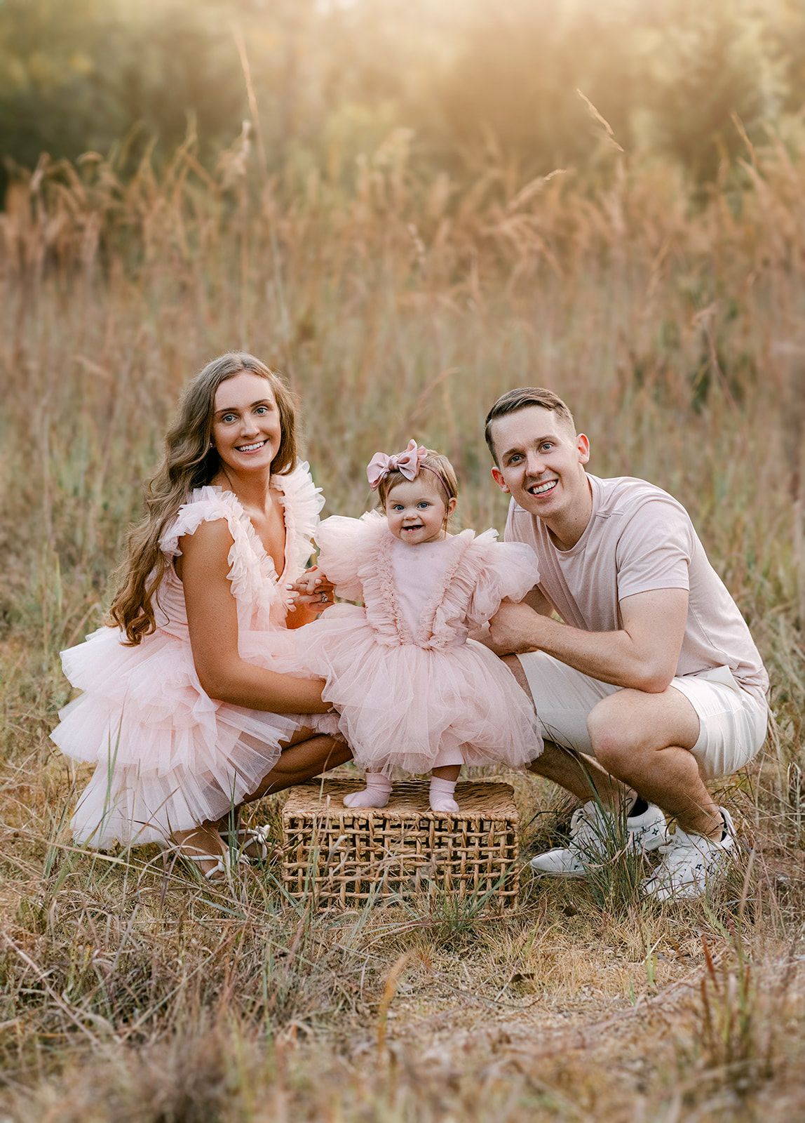 Smiling family with baby girl in pink tutu dress posing in golden grass field at sunset.