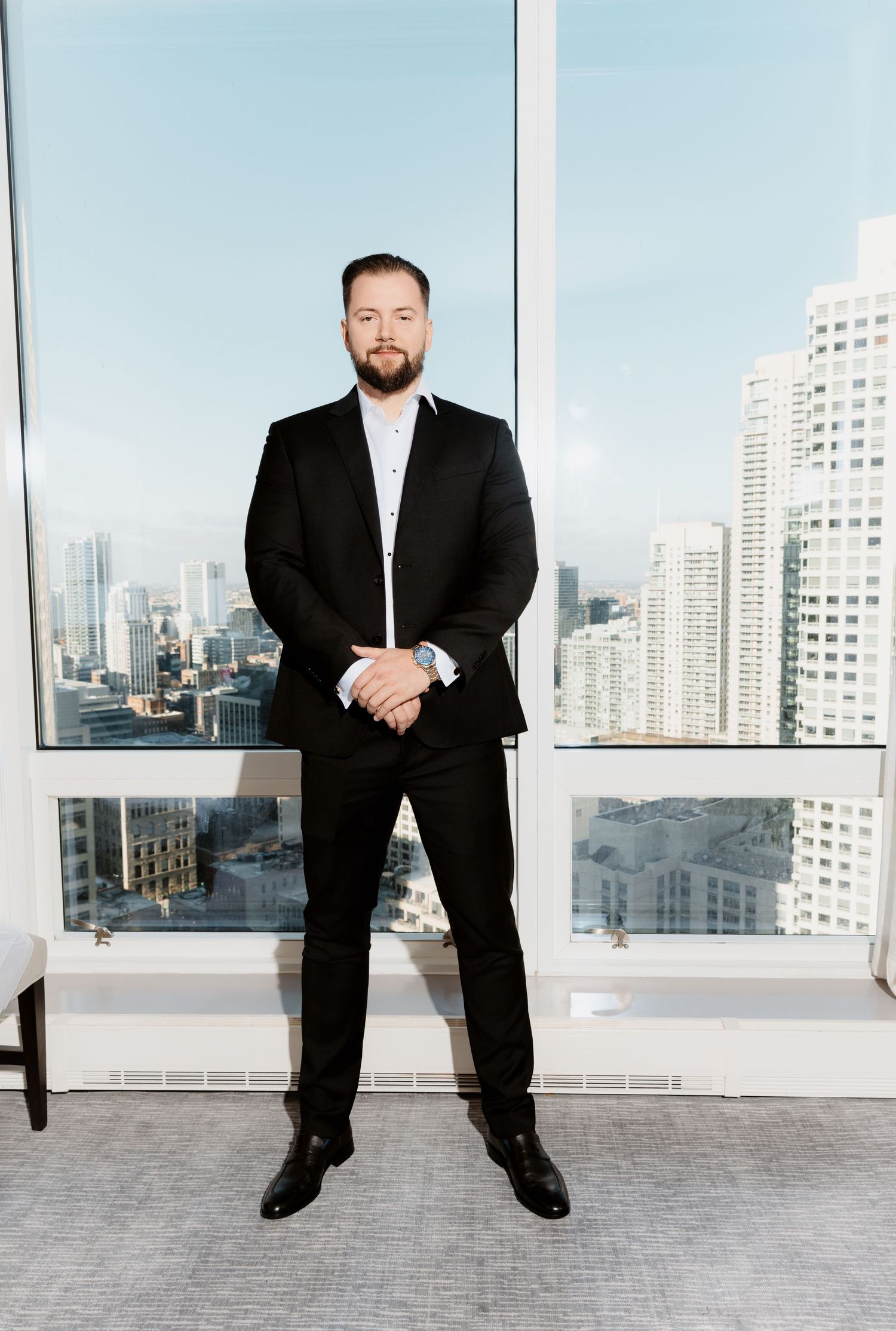 Groom in black suit poses before floor-to-ceiling windows with city skyline backdrop on wedding day.