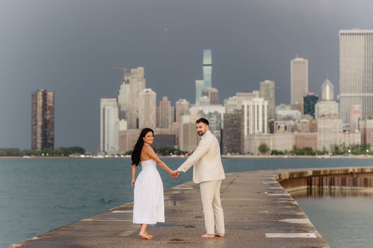 Couple holds hands on Chicago lakefront pier in white dress and beige suit with city skyline backdrop.