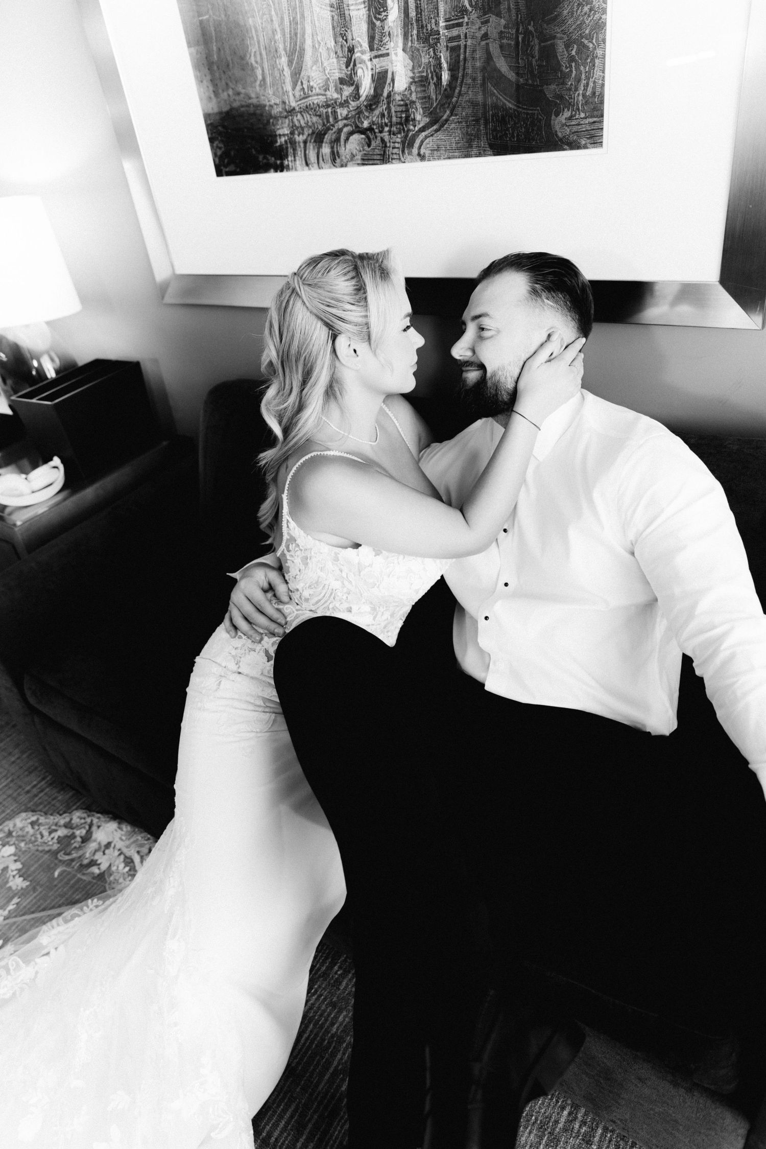 Bride in strapless lace gown and bearded groom share romantic moment on sofa in black and white.