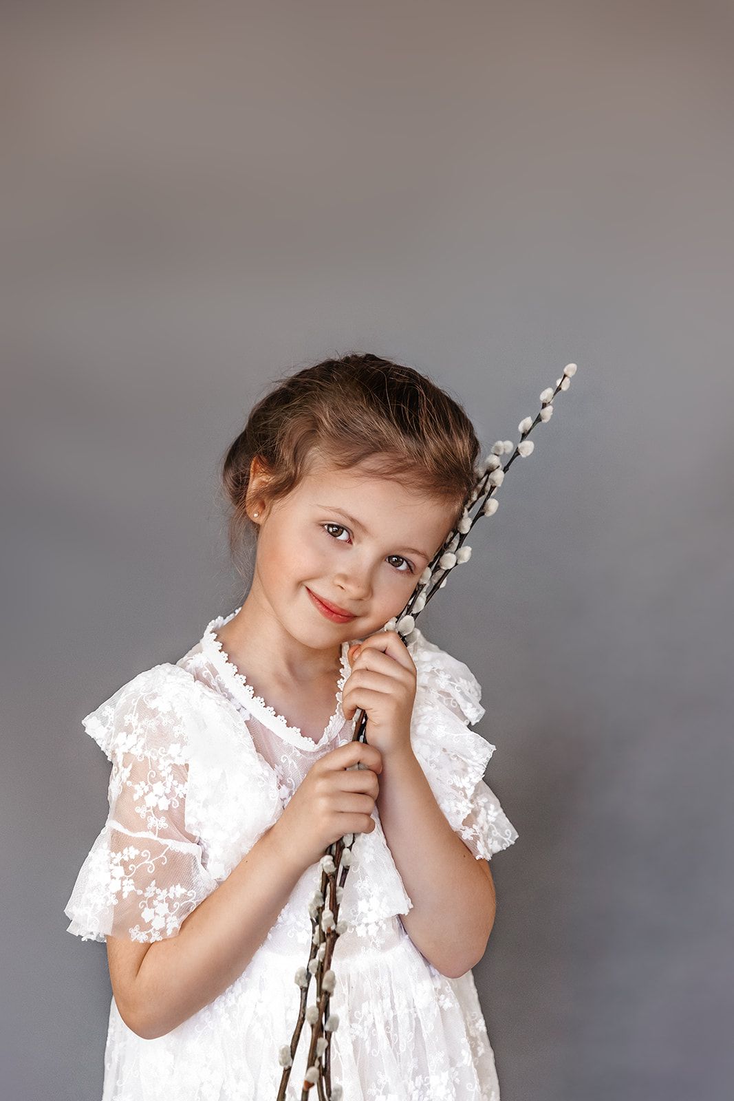 Young girl in white lace dress holding pussy willow branches, smiling against gray background.