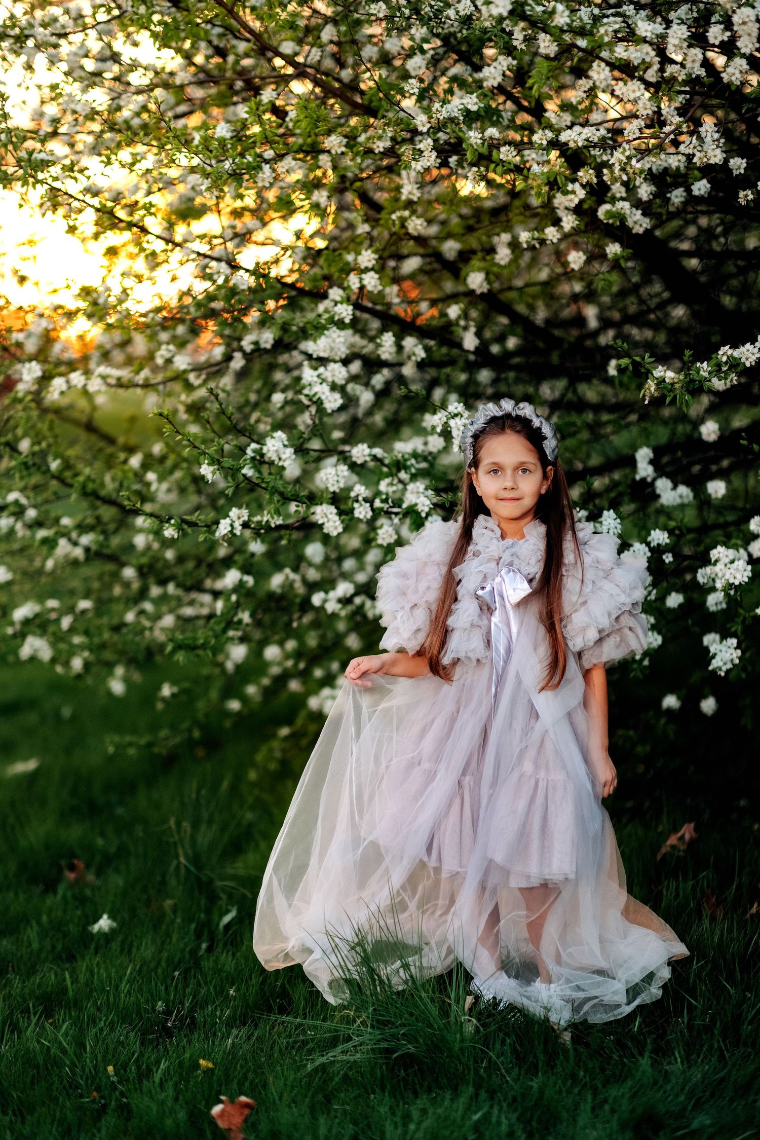 A young girl in a flowing pink princess dress stands gracefully beneath a blooming white cherry blossom tree at sunset.