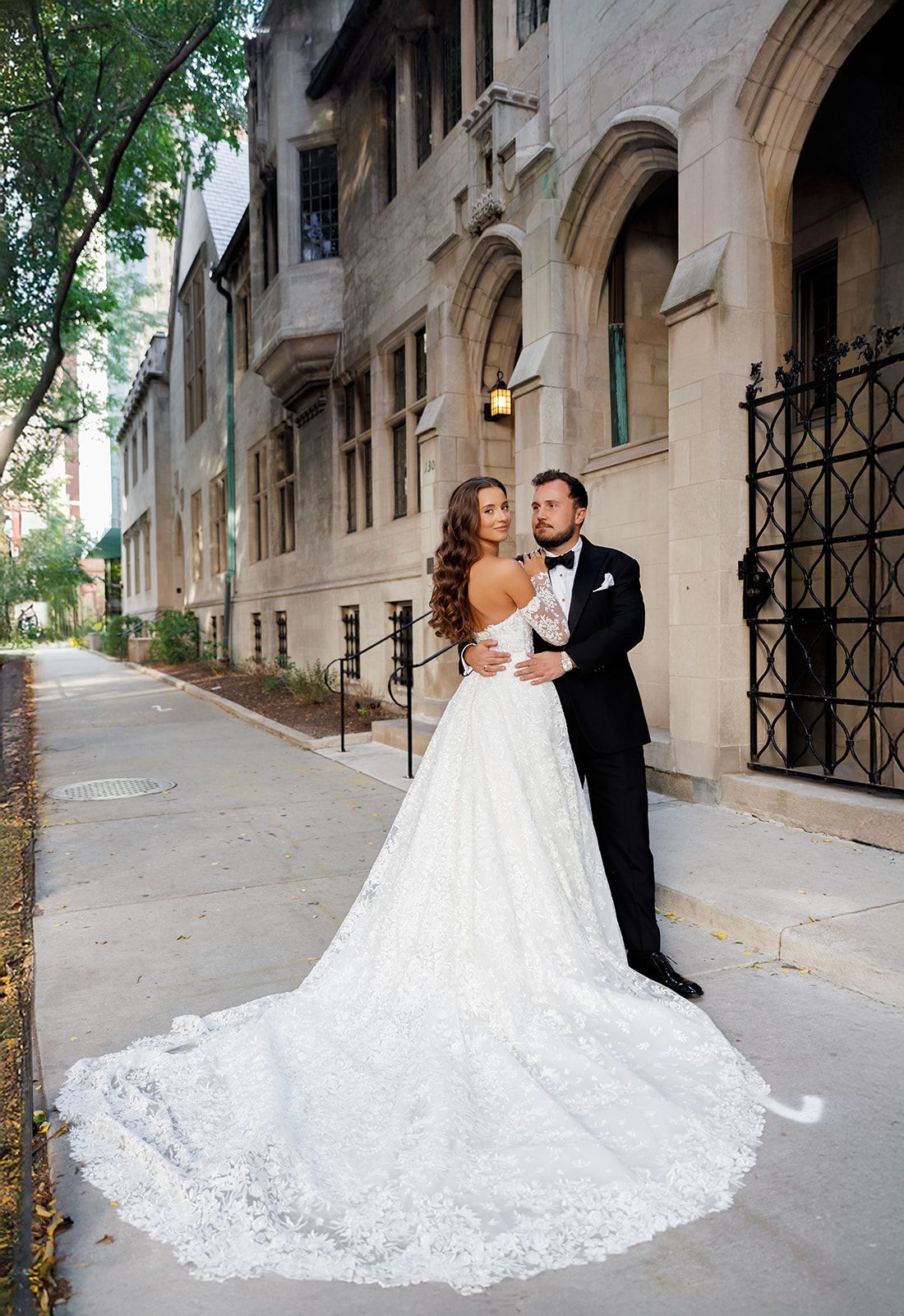 Bride in lace ball gown with long train poses with groom in black tuxedo on urban sidewalk outside stone building.
