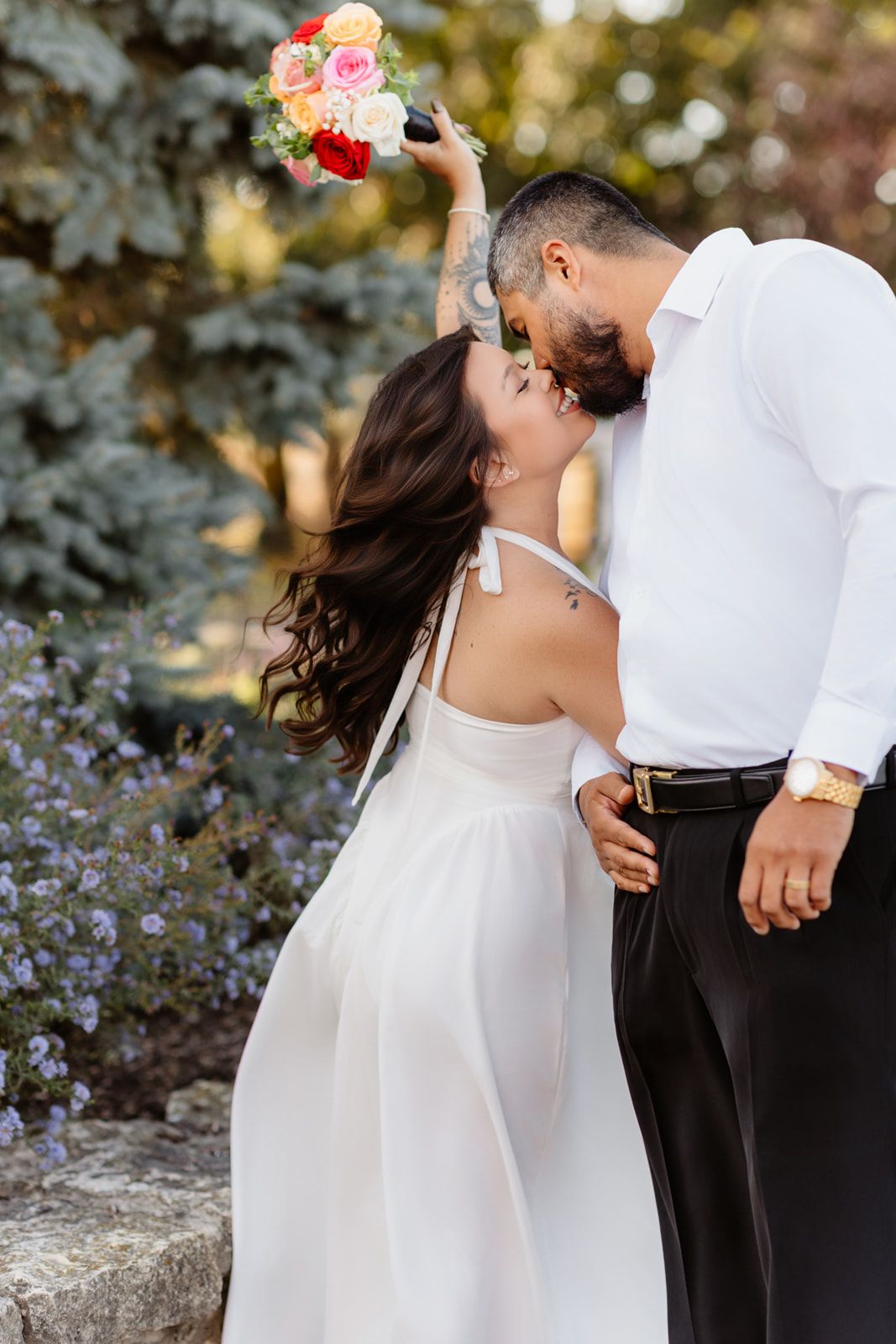Wedding couple kissing in garden, bride in white halter gown raises floral bouquet beside blue spruce tree.