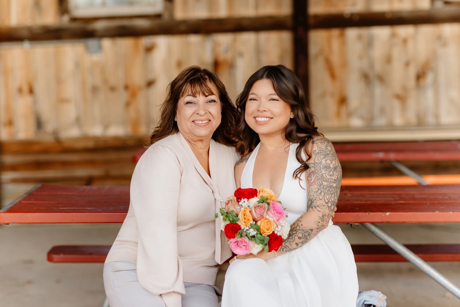 Bride in white gown holding colorful bouquet sits with older woman in blush outfit on red bench in rustic barn setting.