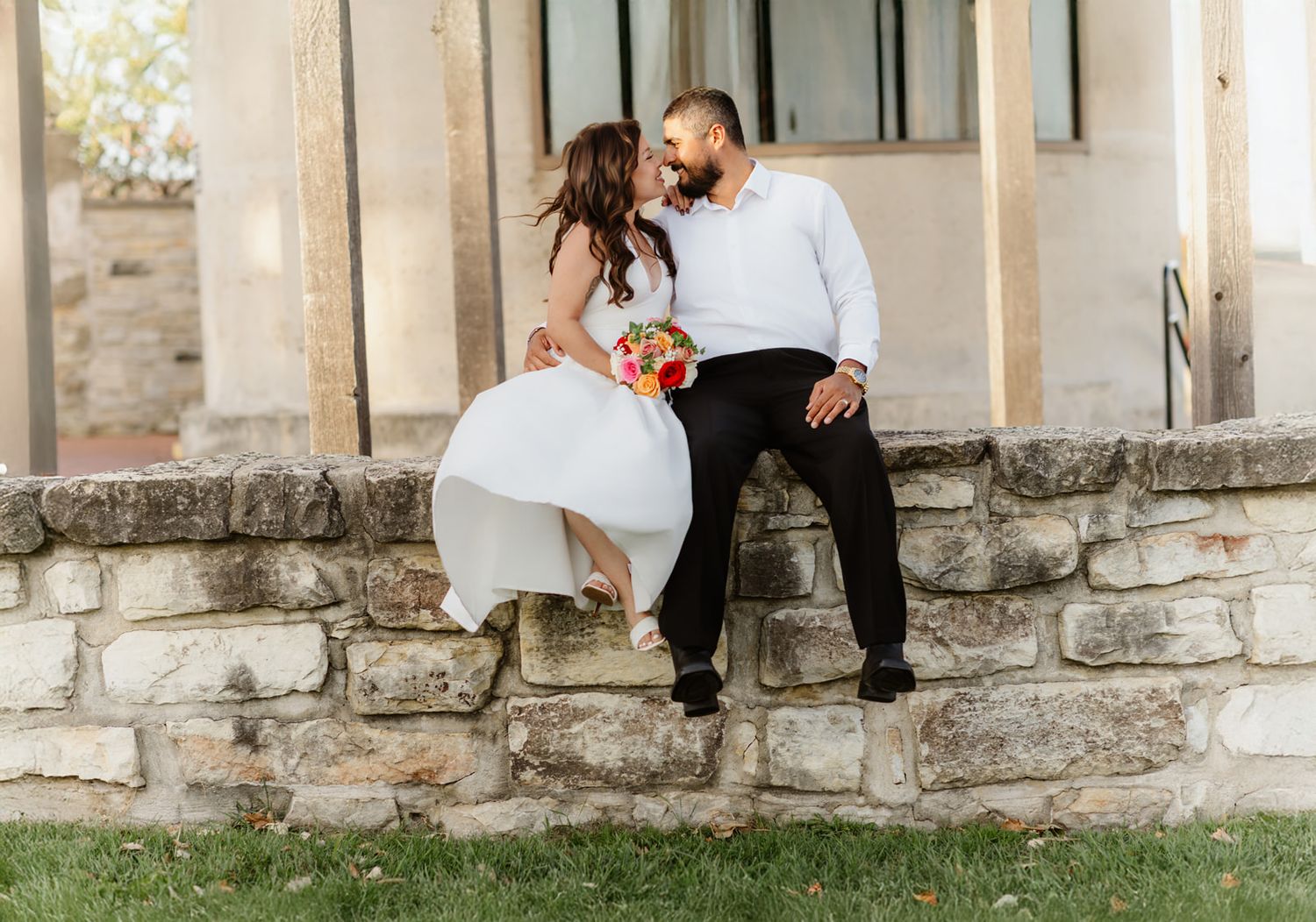 Bride and groom sit on stone wall gazing at each other, bride in white gown holding vibrant bouquet outdoors.