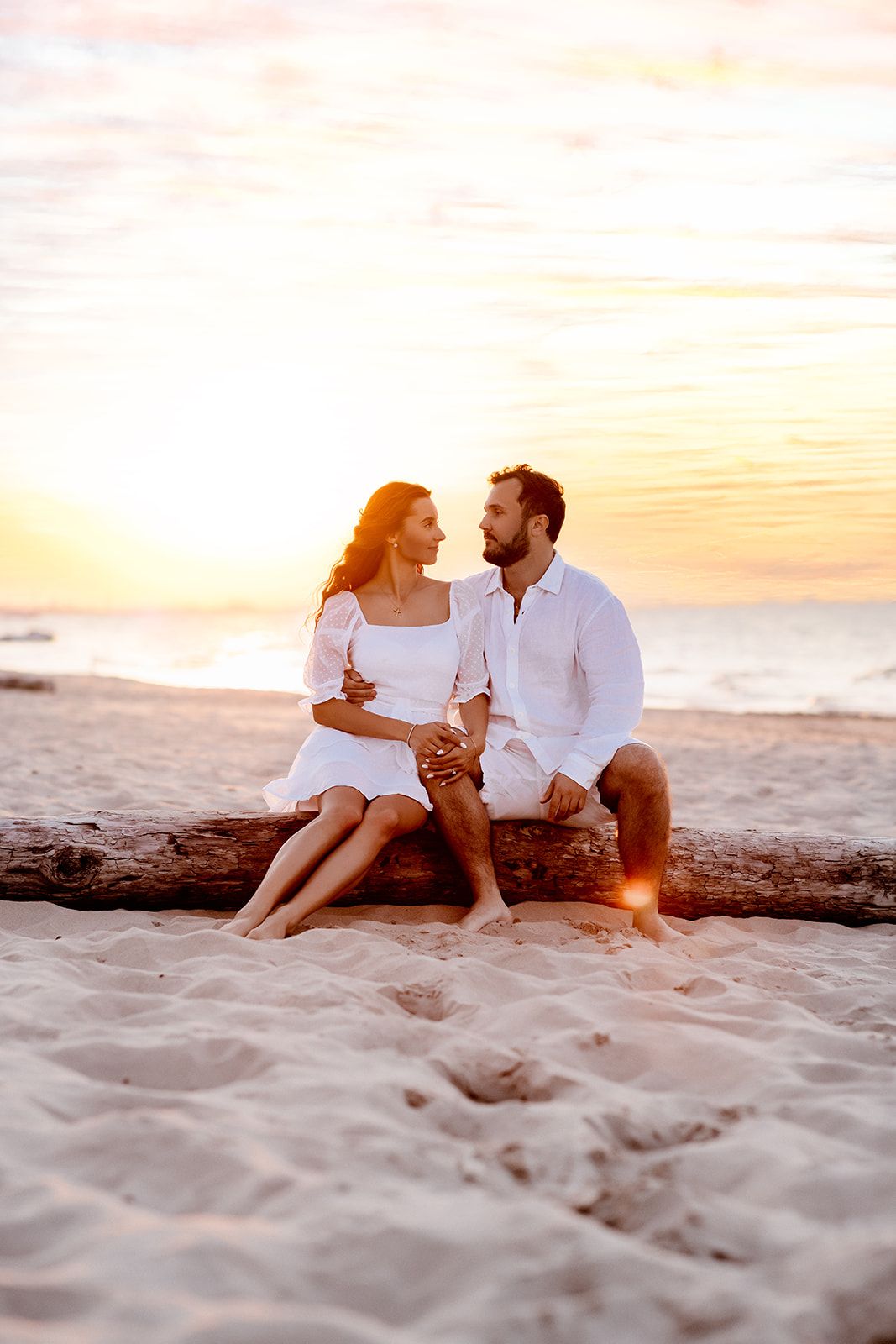 Couple in white outfits sitting on driftwood at a romantic beach sunset, holding hands and gazing at each other.