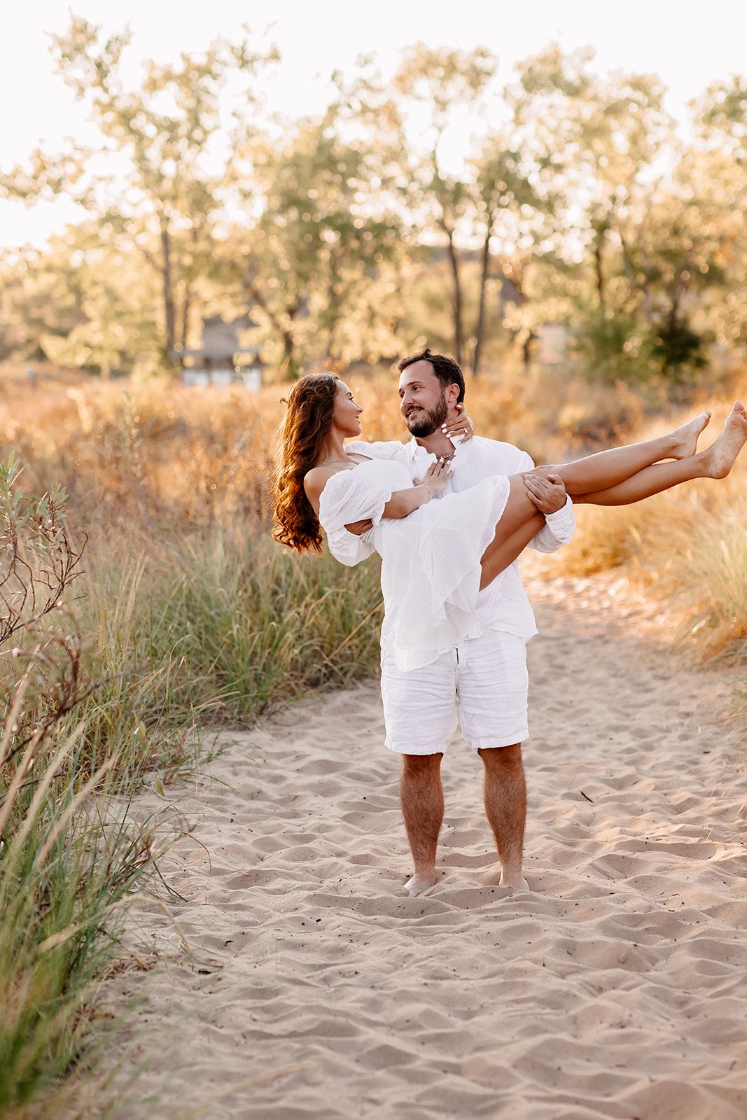 A man in white carries a smiling woman in his arms on a sandy beach path surrounded by golden grass.