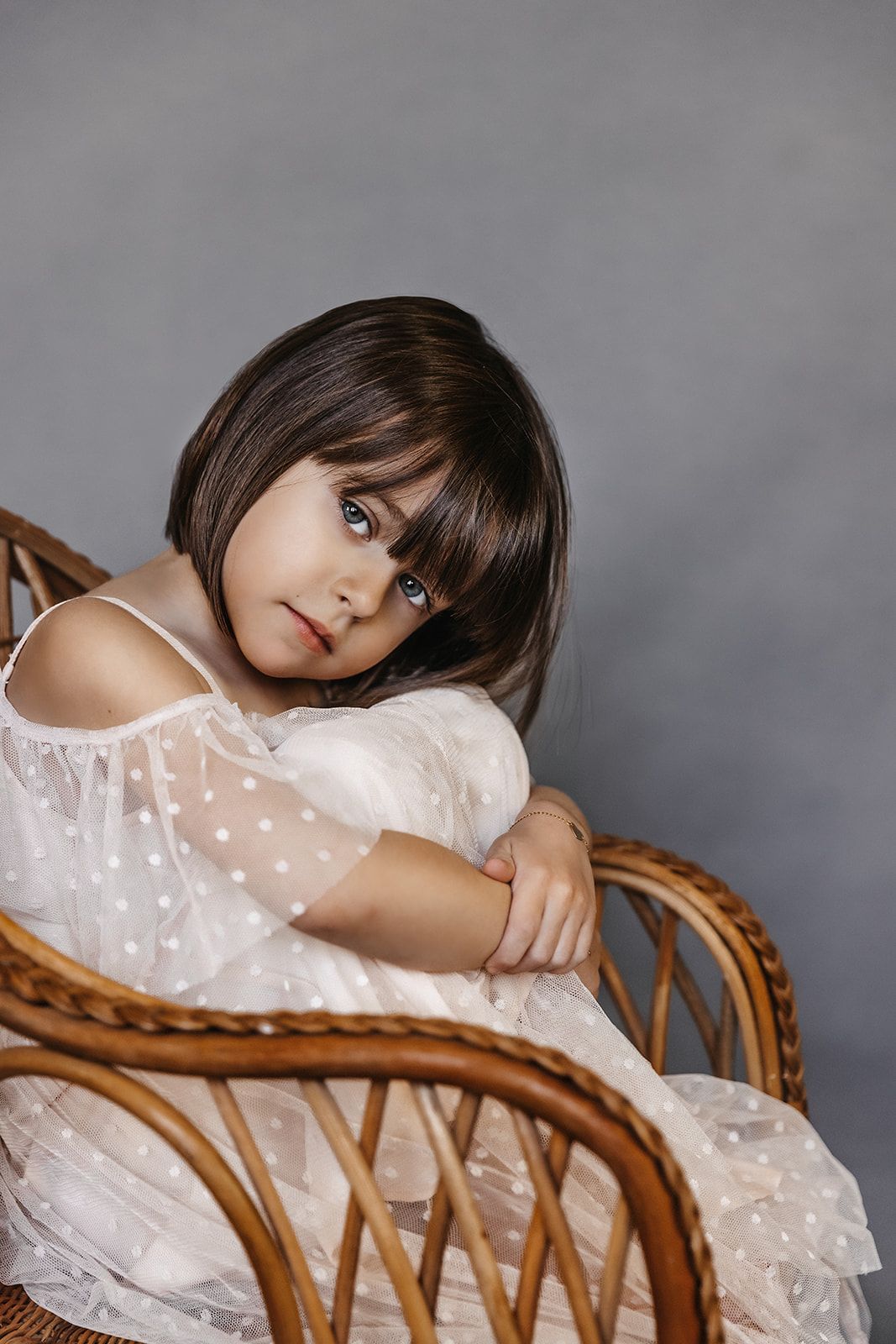 Dark-haired little girl in white polka dot dress sitting in wicker chair against gray background.