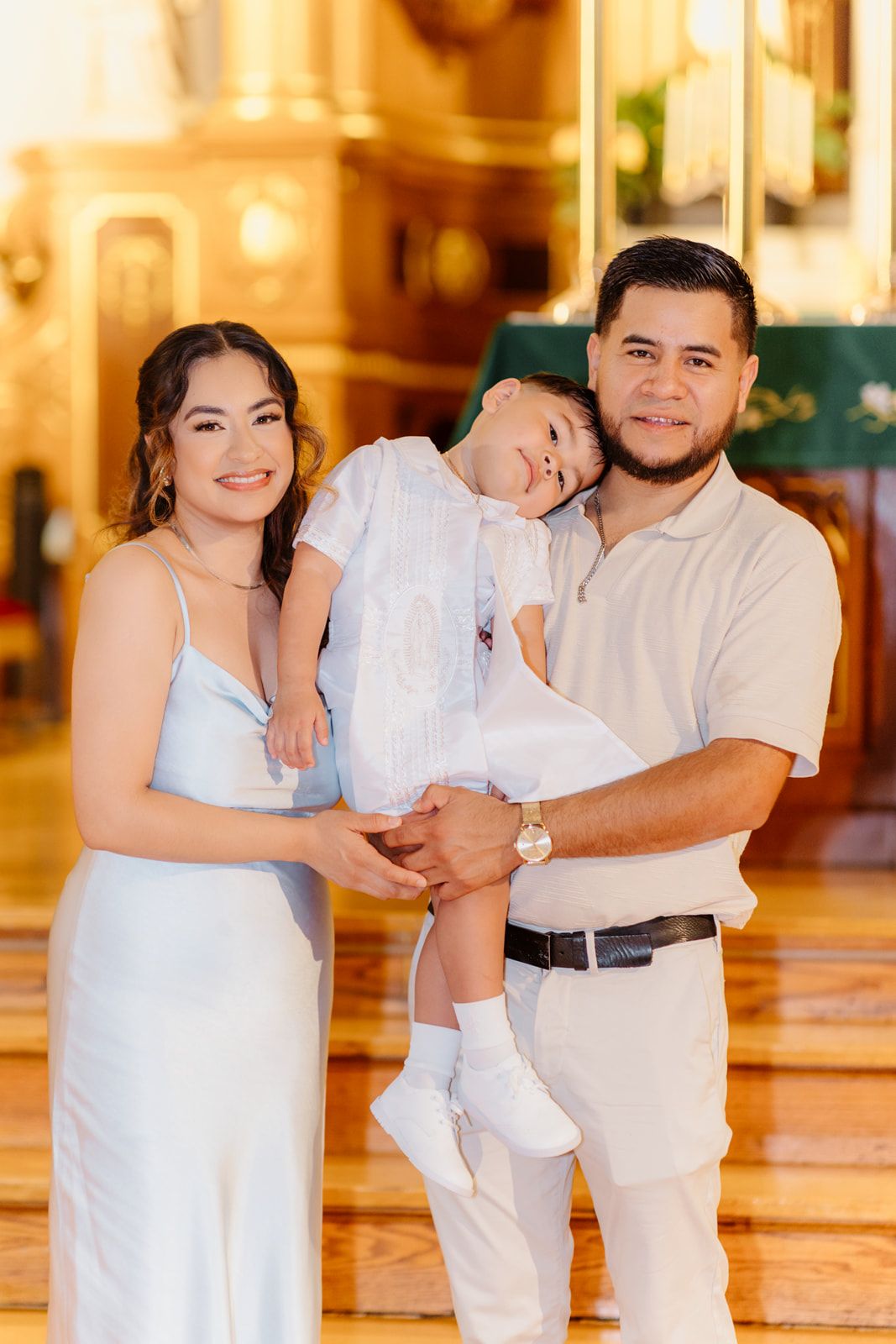 A smiling Hispanic family poses in a church, parents in white holding their baby in a christening outfit.