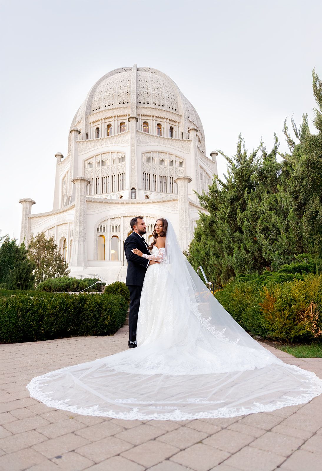 Bride and groom pose before white domed Bahai Temple in Wilmette, Illinois, with sweeping cathedral veil trailing on stone plaza.