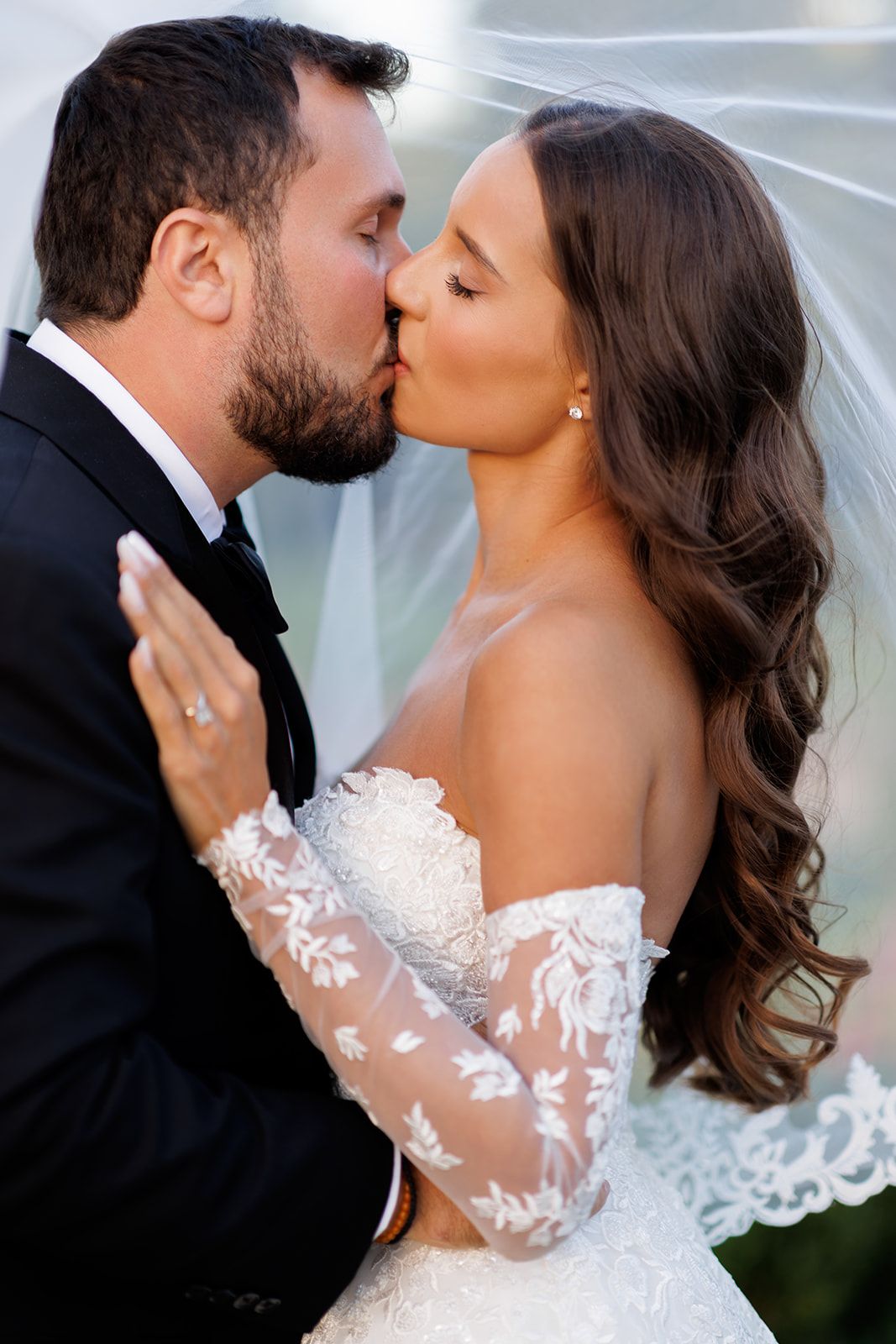 Bride and groom share romantic kiss under veil, bride in off-shoulder lace gown with floral embroidery.
