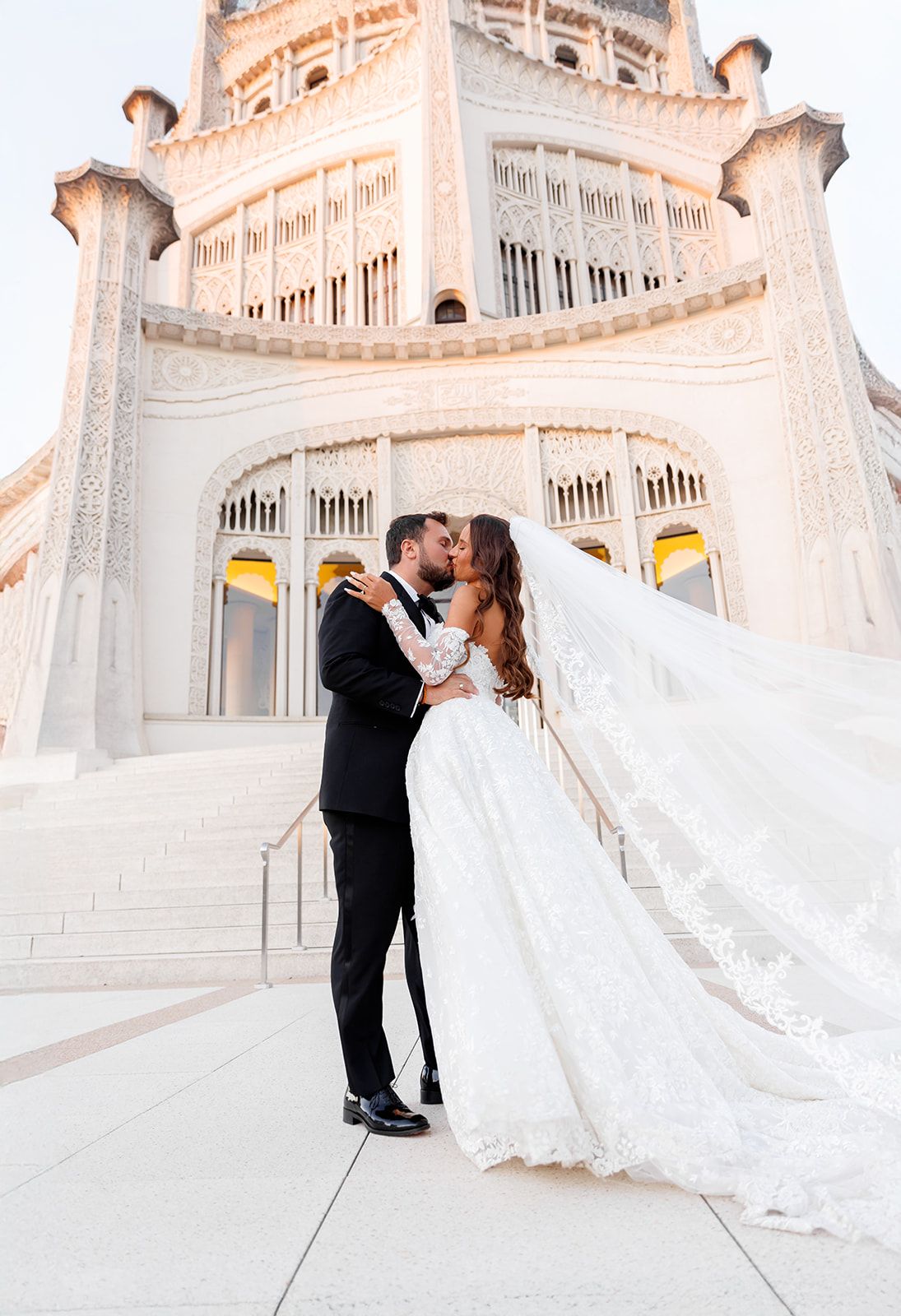 Bride and groom share a romantic kiss in front of an ornate white temple, her lace gown and veil flowing gracefully.