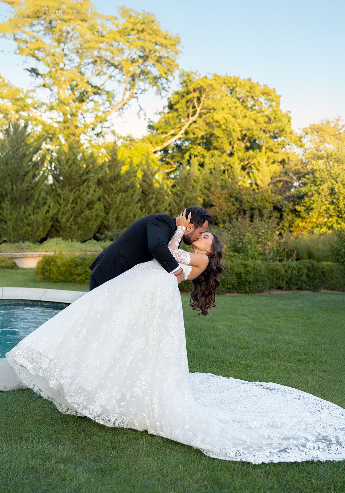 Groom dramatically dips bride for a kiss outdoors, her lace wedding gown train spread across green grass.