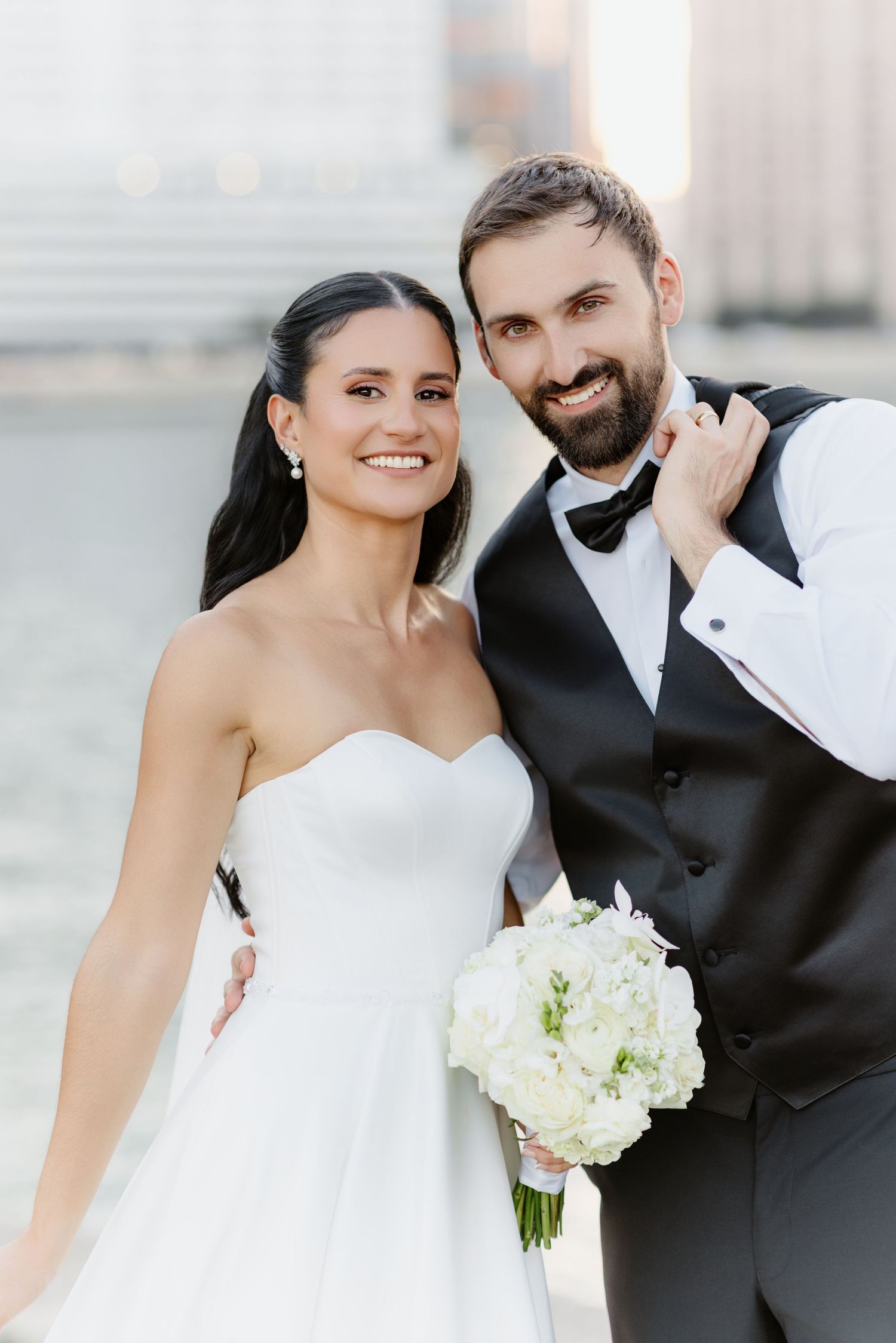 Happy bride in strapless white gown and bearded groom in black vest and bow tie pose with white bouquet.