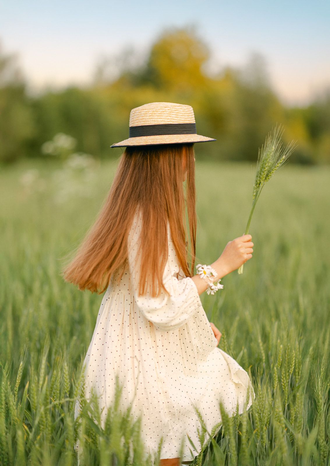 Back view of a girl in straw hat and white dress holding wheat stalks in a green summer field.