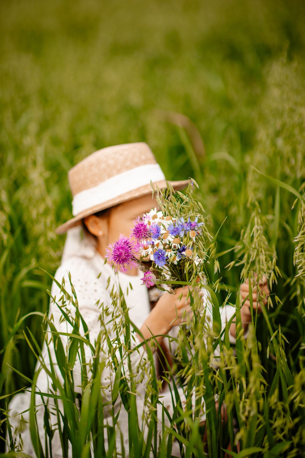 Child in straw hat holding colorful wildflower bouquet while sitting in tall green grass field.