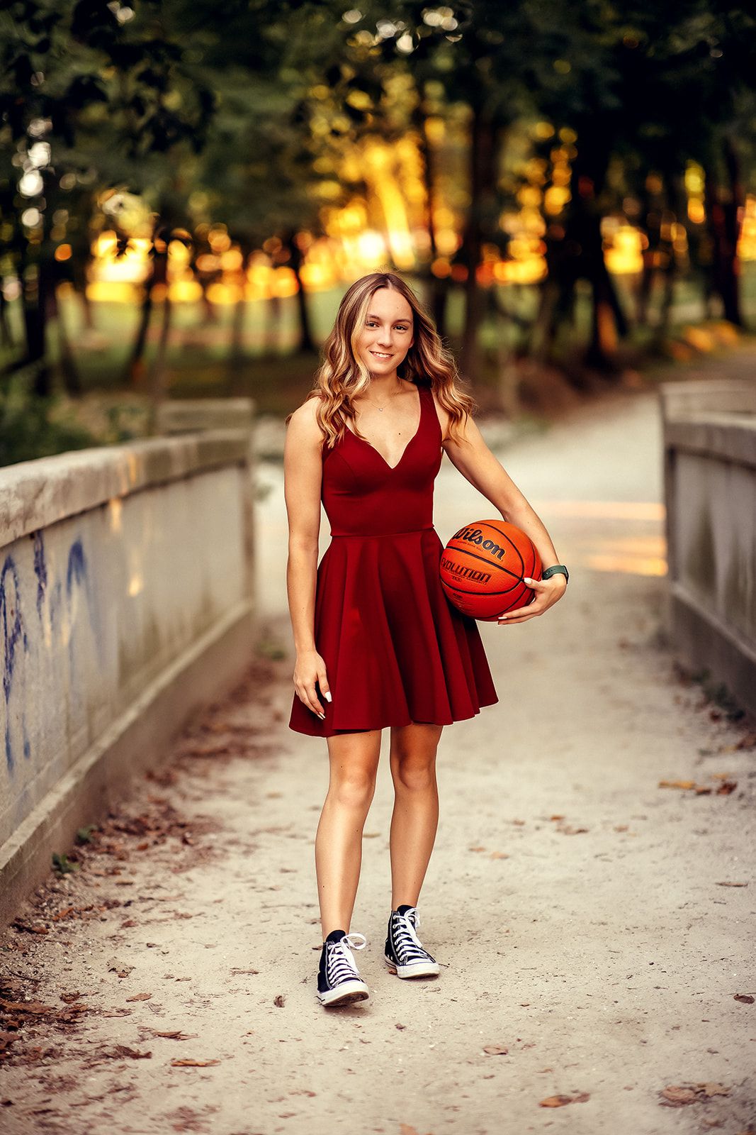 Young woman in red dress and Converse sneakers holds a basketball on a bridge path at sunset.