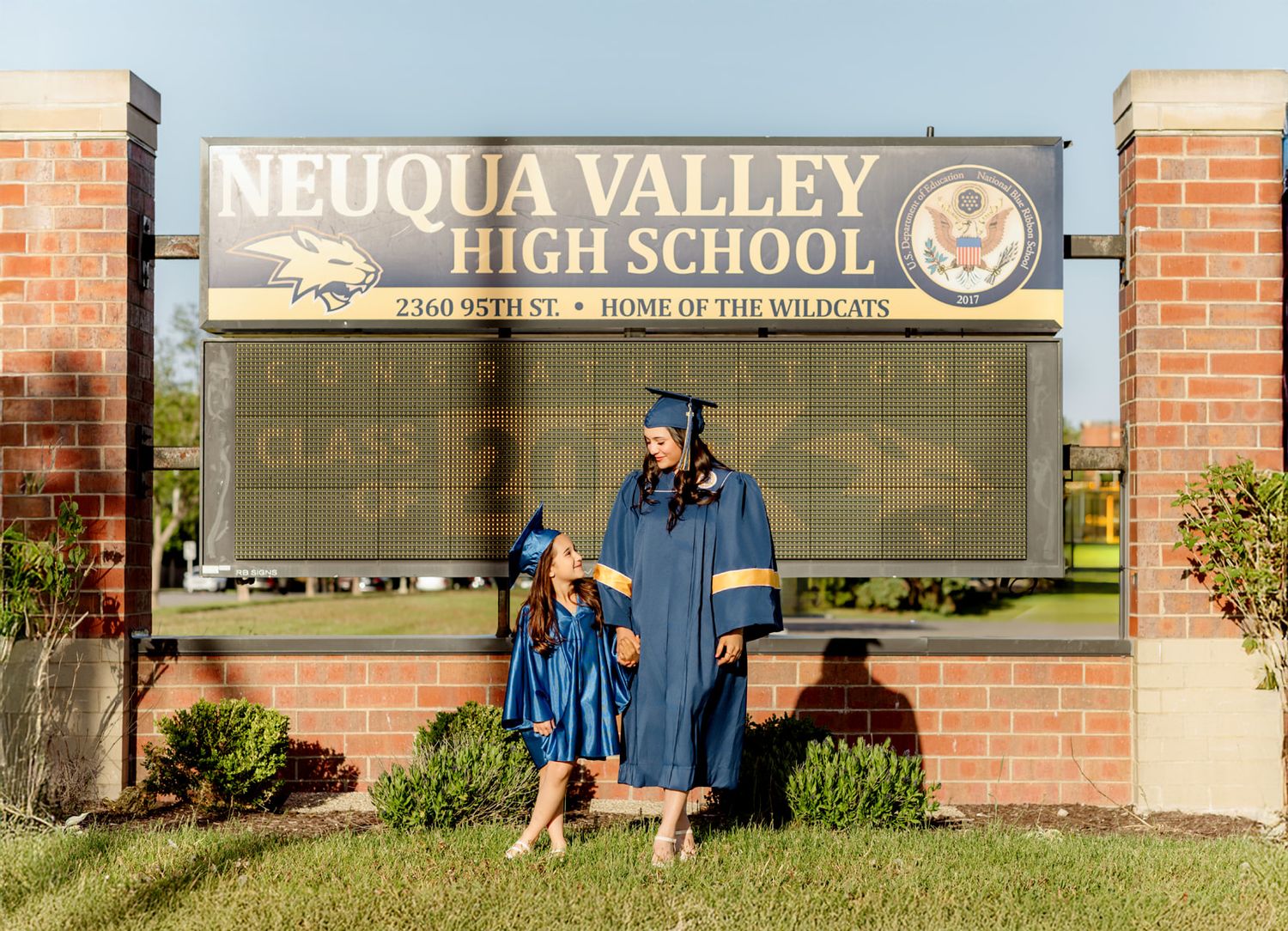 Two graduates in blue caps and gowns posing in front of Neuqua Valley High School sign.
